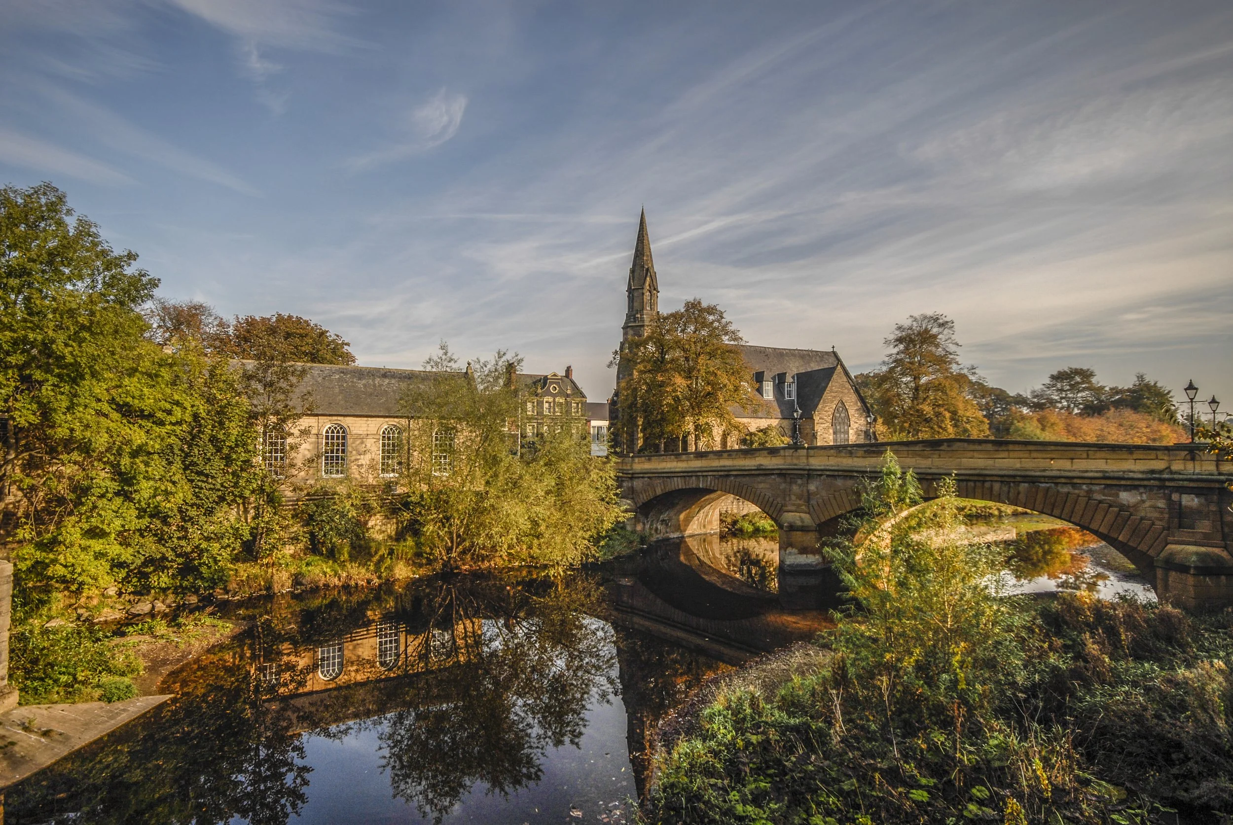 A church with a tall steeple surrounded by trees with colorful autumn leaves, a stone bridge over a river, and a blue sky with wispy clouds.