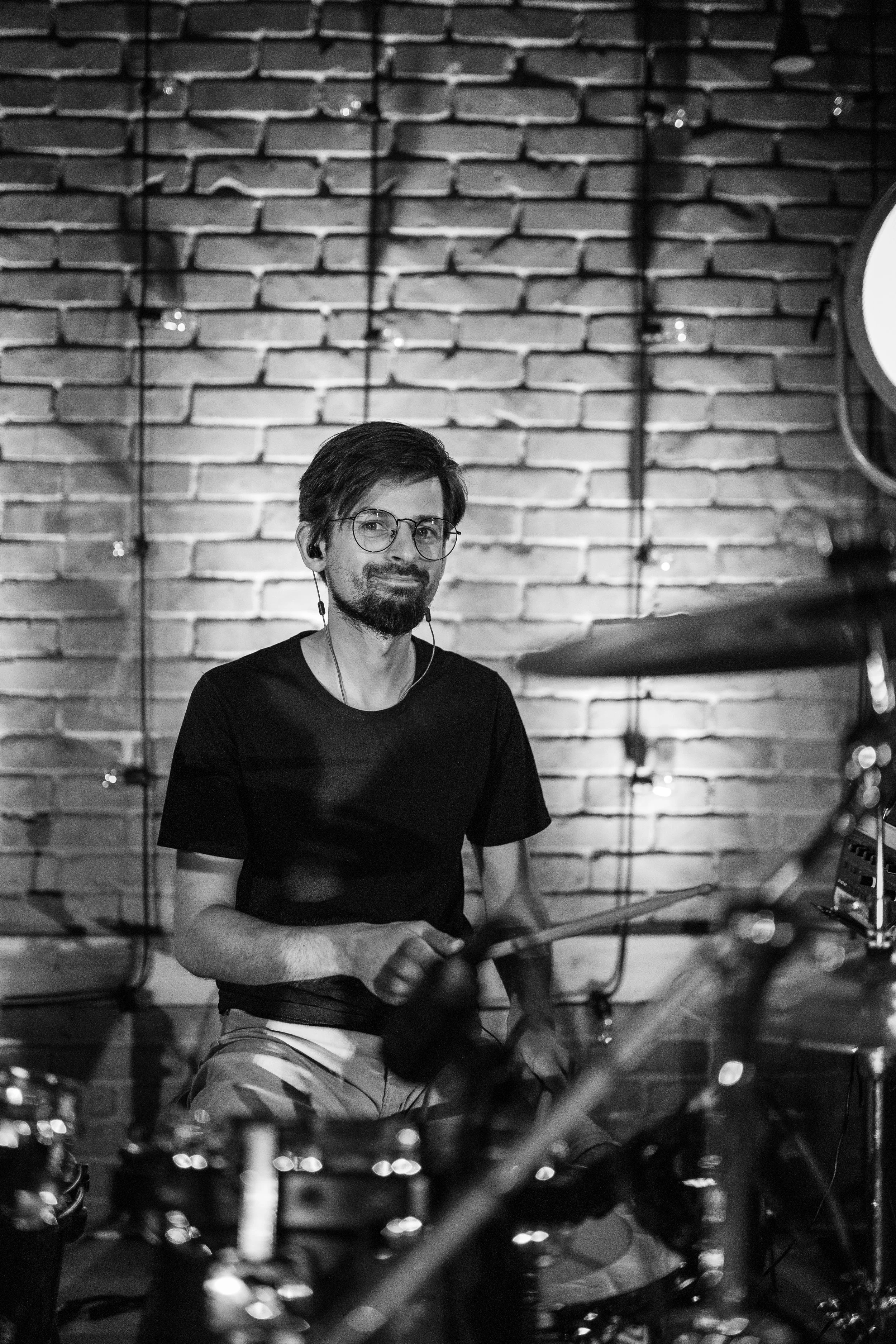 A man with glasses and facial hair sitting in front of a brick wall, wearing a black t-shirt and striped shorts, surrounded by musical instruments, possibly in a recording studio.