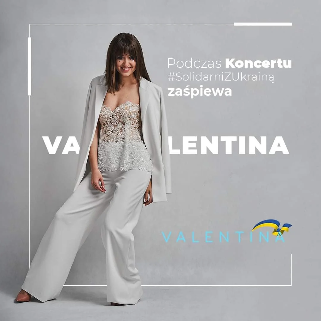 Woman in white pants, a beige lace top, and a white blazer standing against a gray background with Polish text and a logo, promoting a concert for Ukraine solidarity.