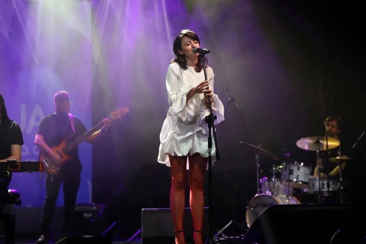 Female singer performing on stage with a microphone, accompanied by a band including a bassist and drummer, under colorful stage lights.