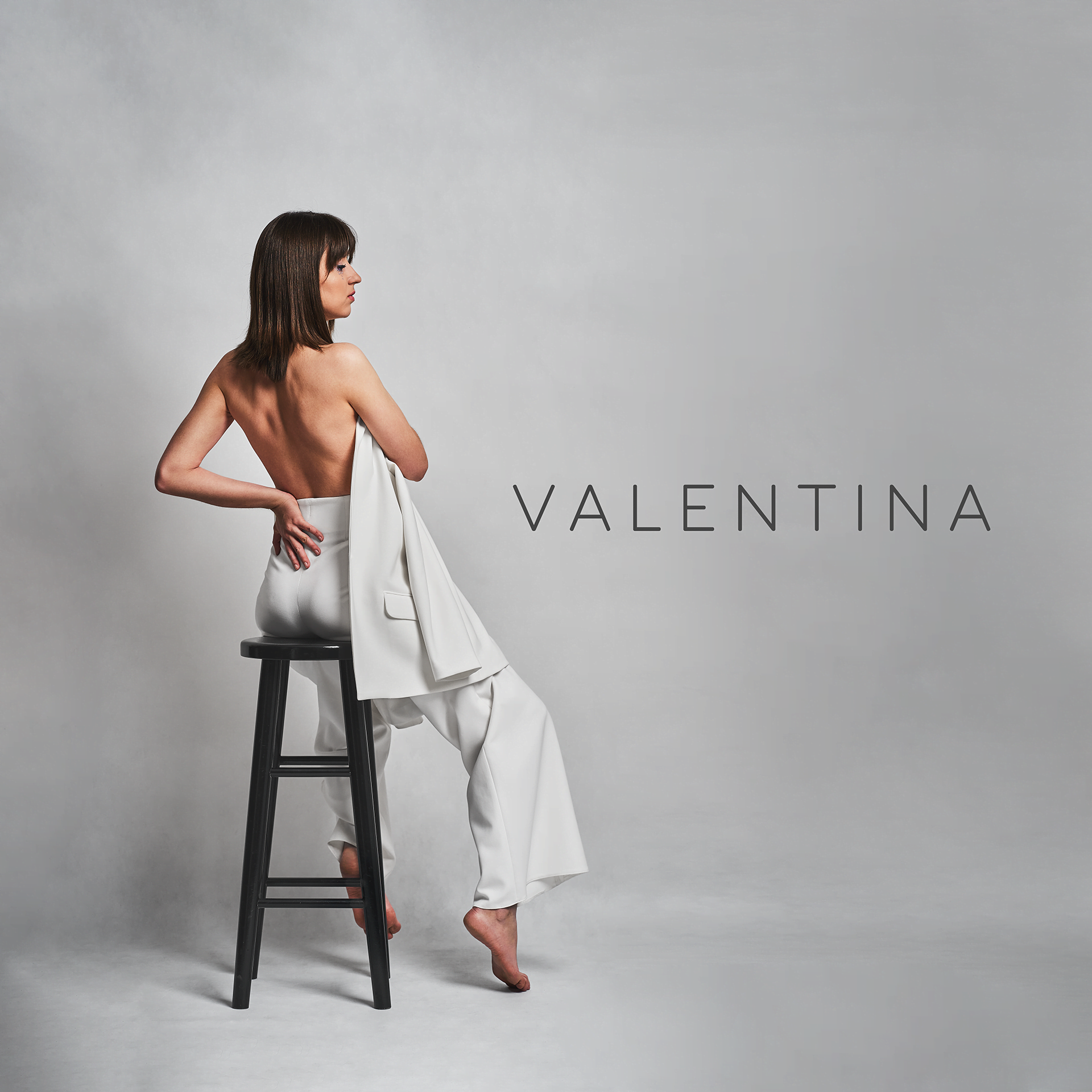 A woman with dark brown hair in a bob style, sitting on a black stool with her back partly covered by a white blazer draped over her shoulder, facing sideways with her eyes closed, in front of a plain light gray background.