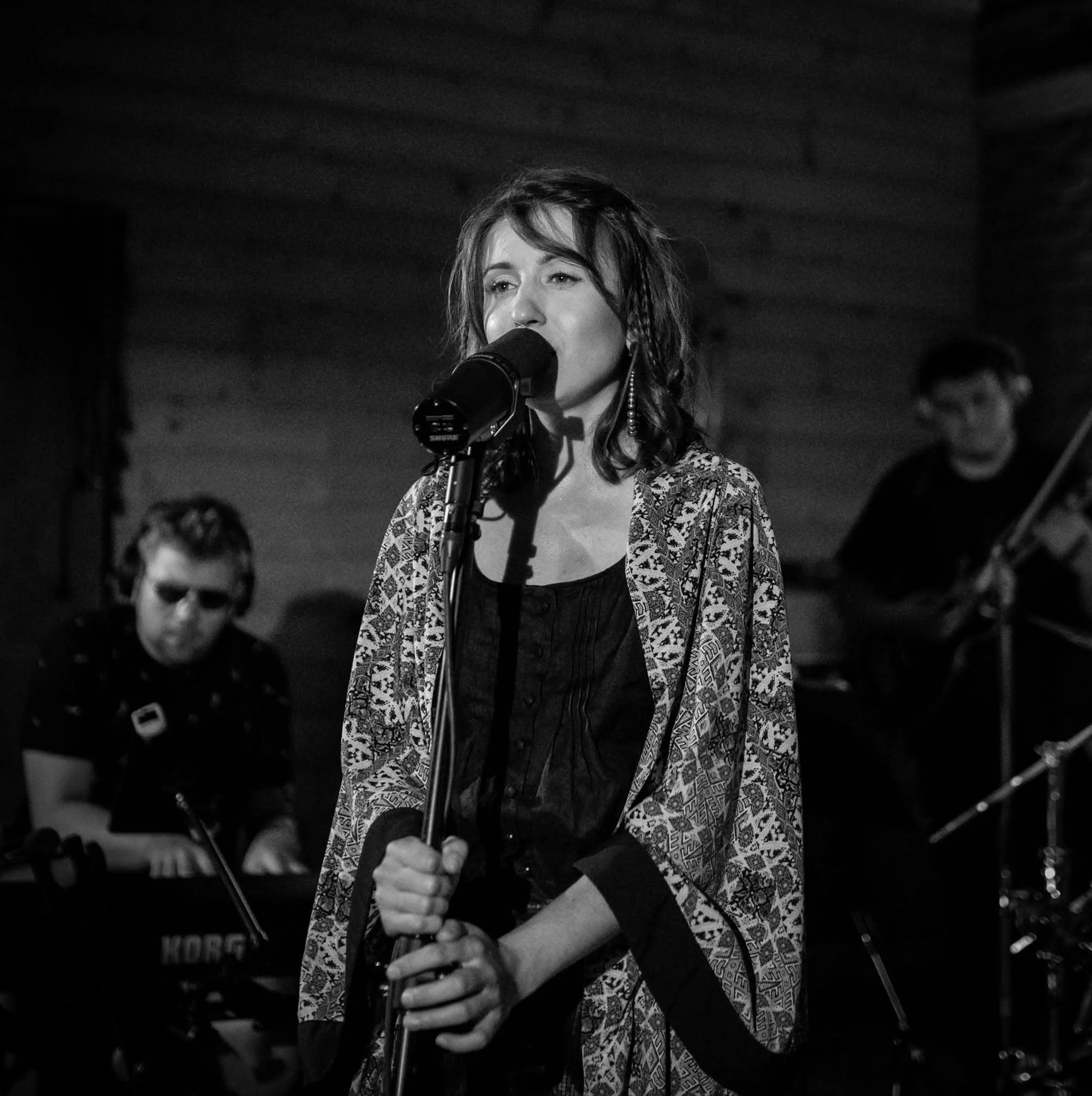Black-and-white image of a woman singing into a microphone during a performance, with a musician playing a keyboard in the background.