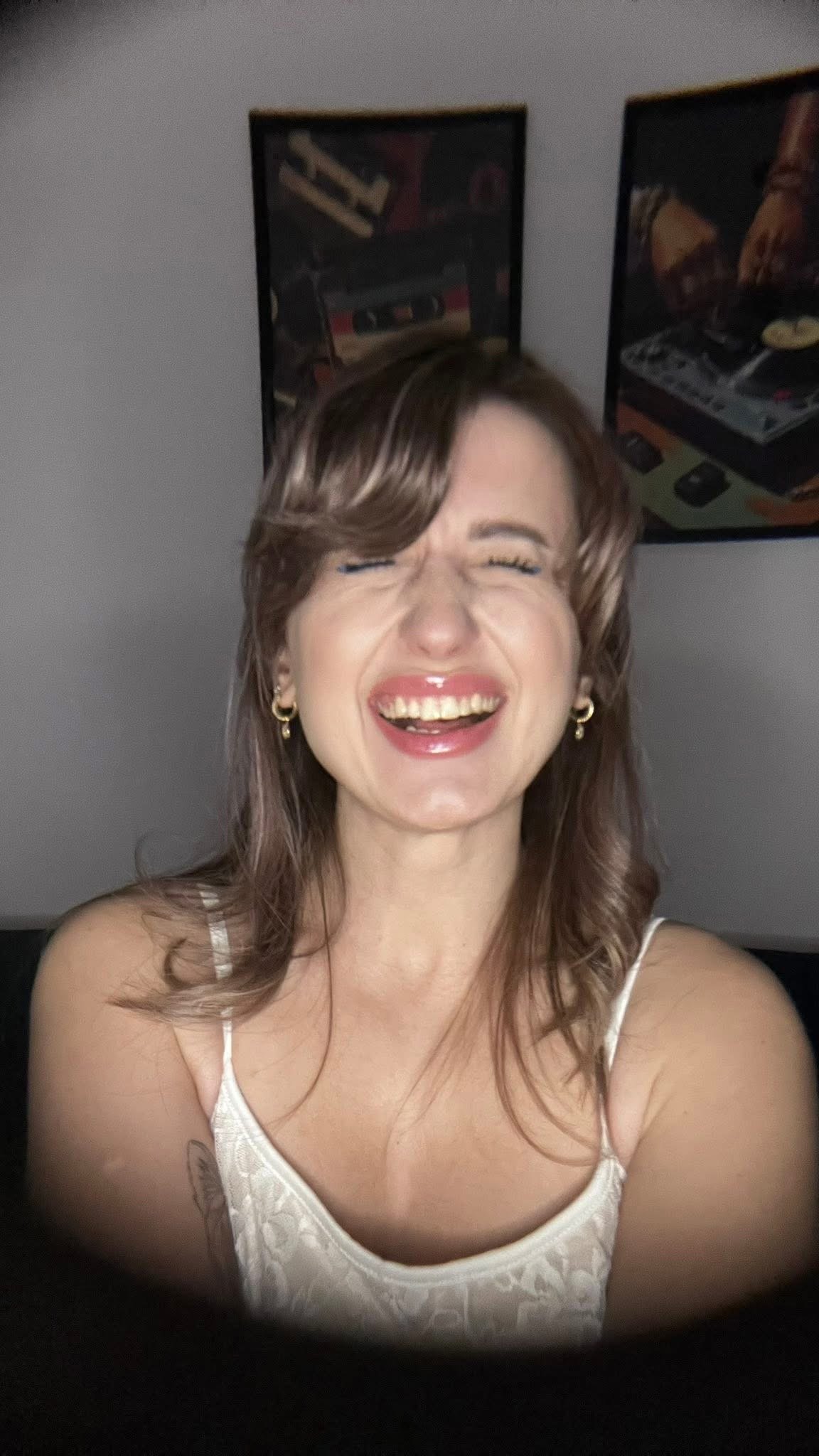 A woman with brown hair smiling and showing her teeth, wearing earrings and a white spaghetti strap top, with two framed pictures of vintage arcade games hanging on the wall behind her.