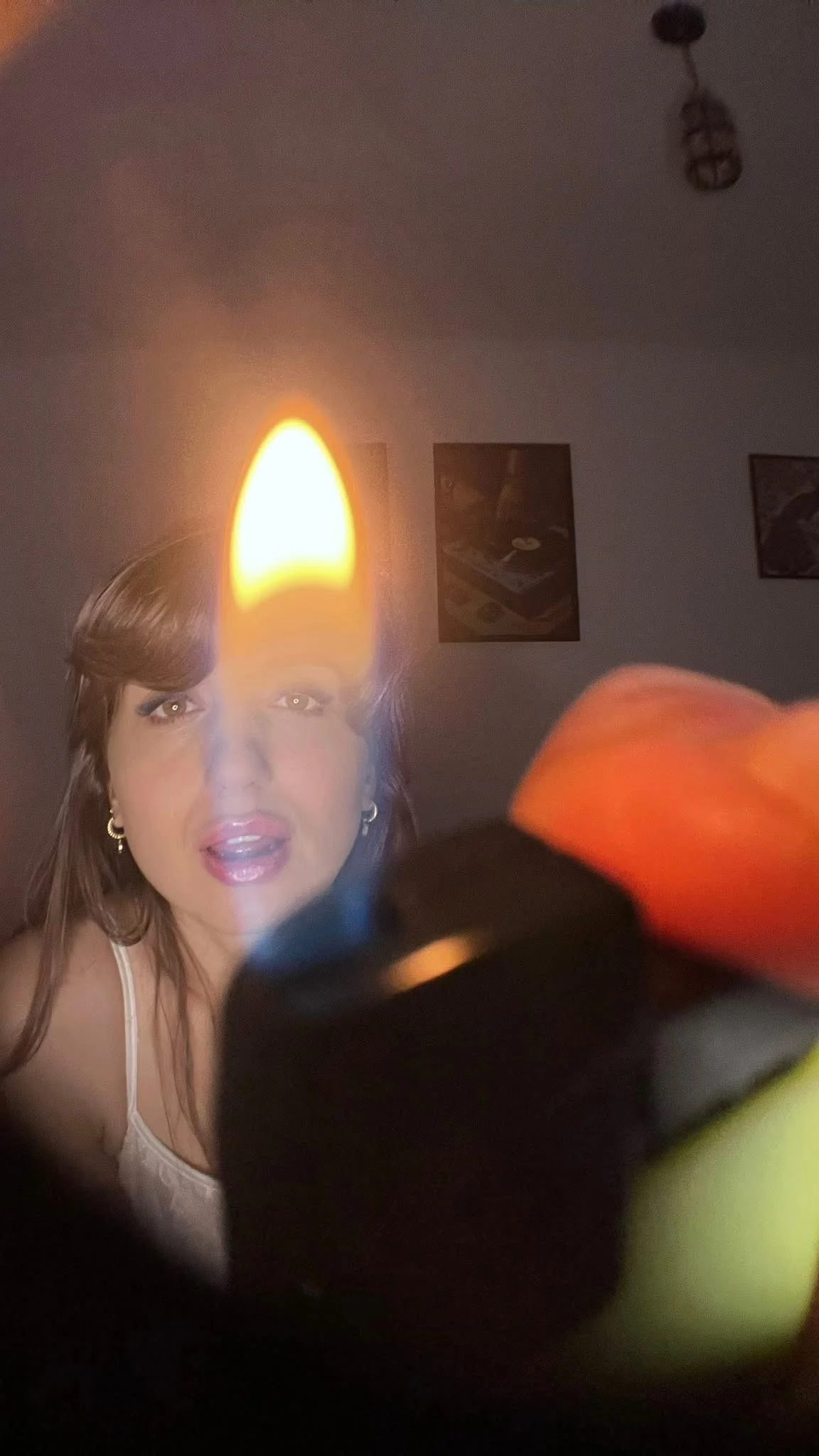 A woman taking a selfie with a lit candle in the foreground, partially covering her face. The background shows a ceiling light fixture, framed pictures on the wall, and a dark room.