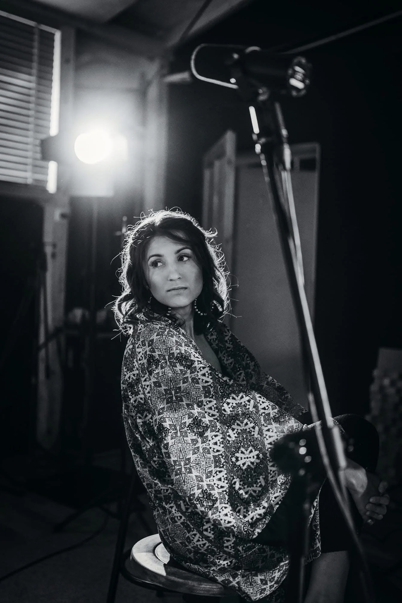 A woman with dark, wavy hair sits on a wooden chair, in a room with dark walls, wearing a patterned loose-fitting garment. She is holding an acoustic guitar and looking off to the side. The room is dimly lit with a bright light source to the left, cr