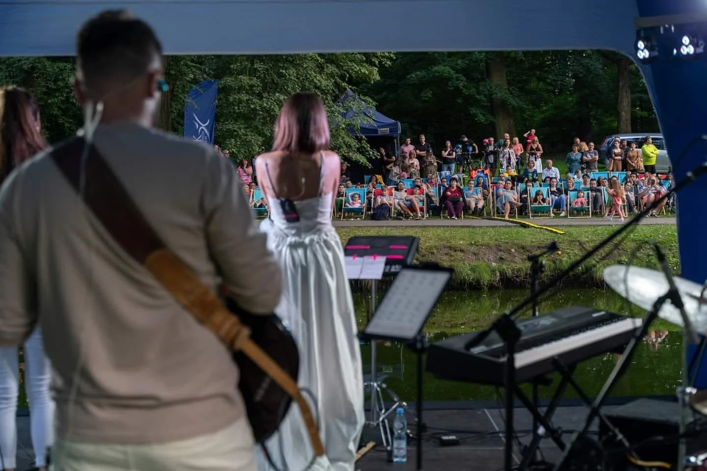 Musicians performing at an outdoor concert by a pond, with an audience seated in chairs and standing among trees in the background.