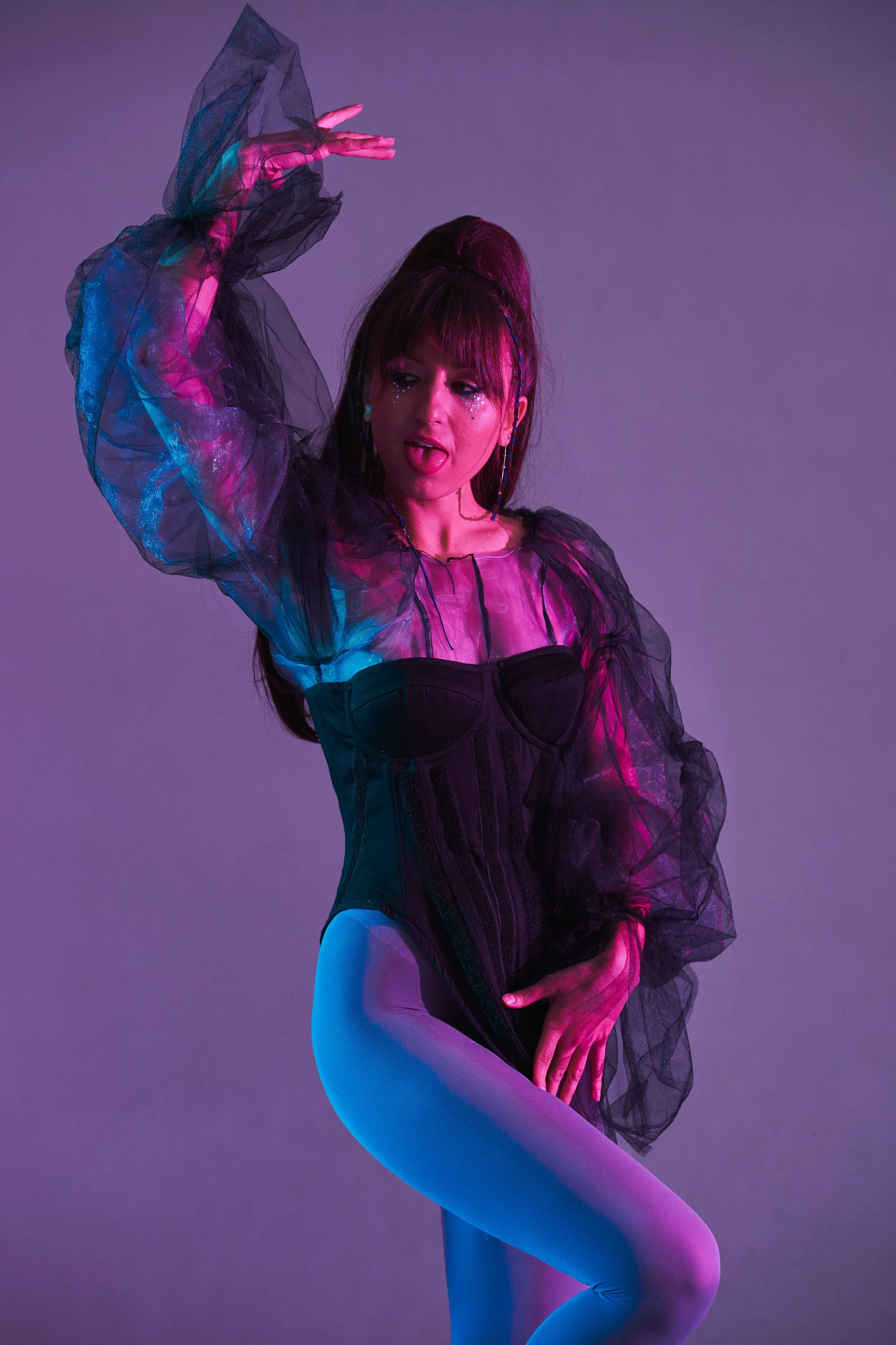 A woman in colorful, shiny dancewear with sheer, ruffled sleeves and light blue tights is posing against a purple background. She has dark hair with bangs and streaked makeup, with sparkles around her eyes.