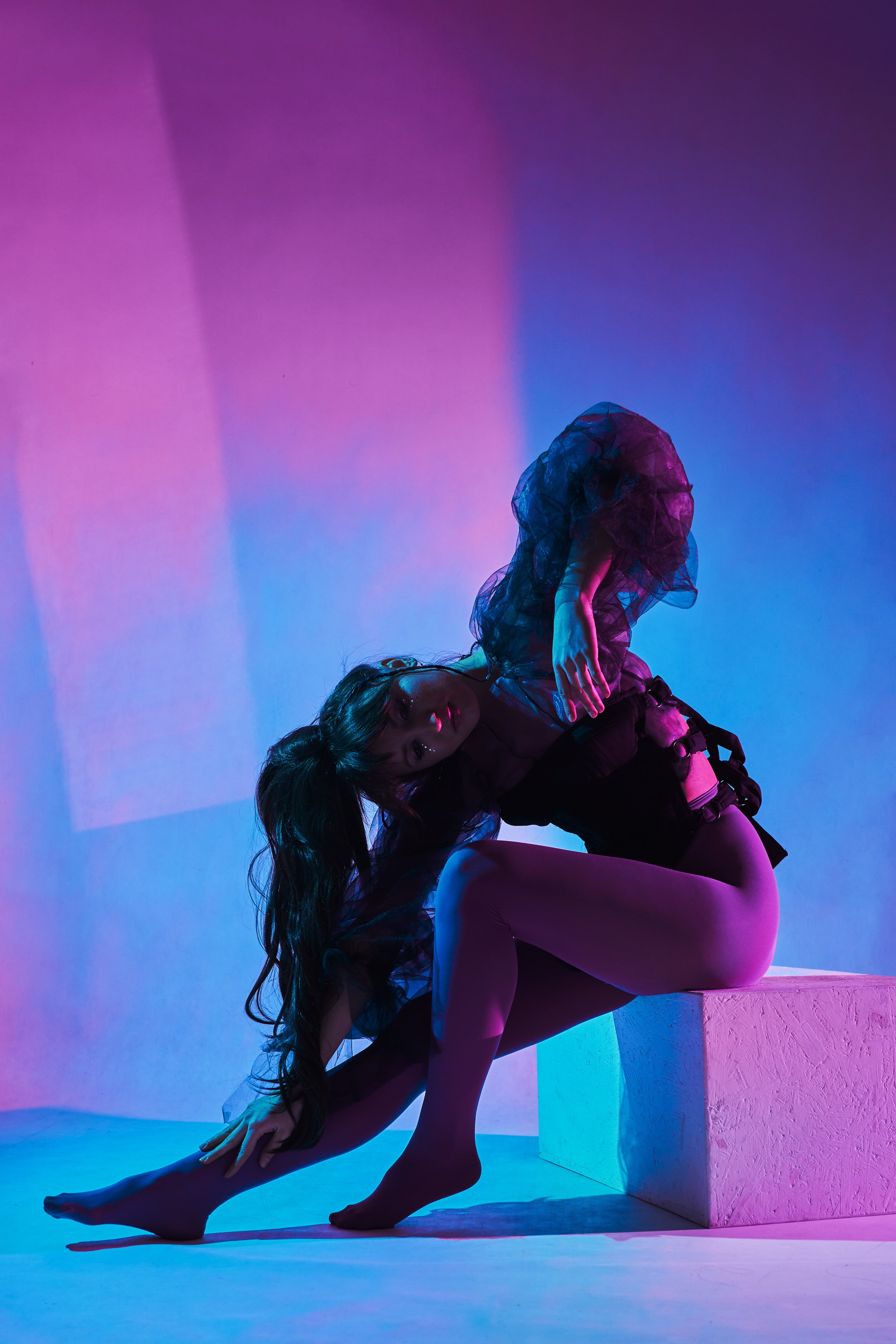 Two women engaged in a dance pose with colorful purple, pink, and blue lighting, one bending backwards on a white block, the other supporting her.