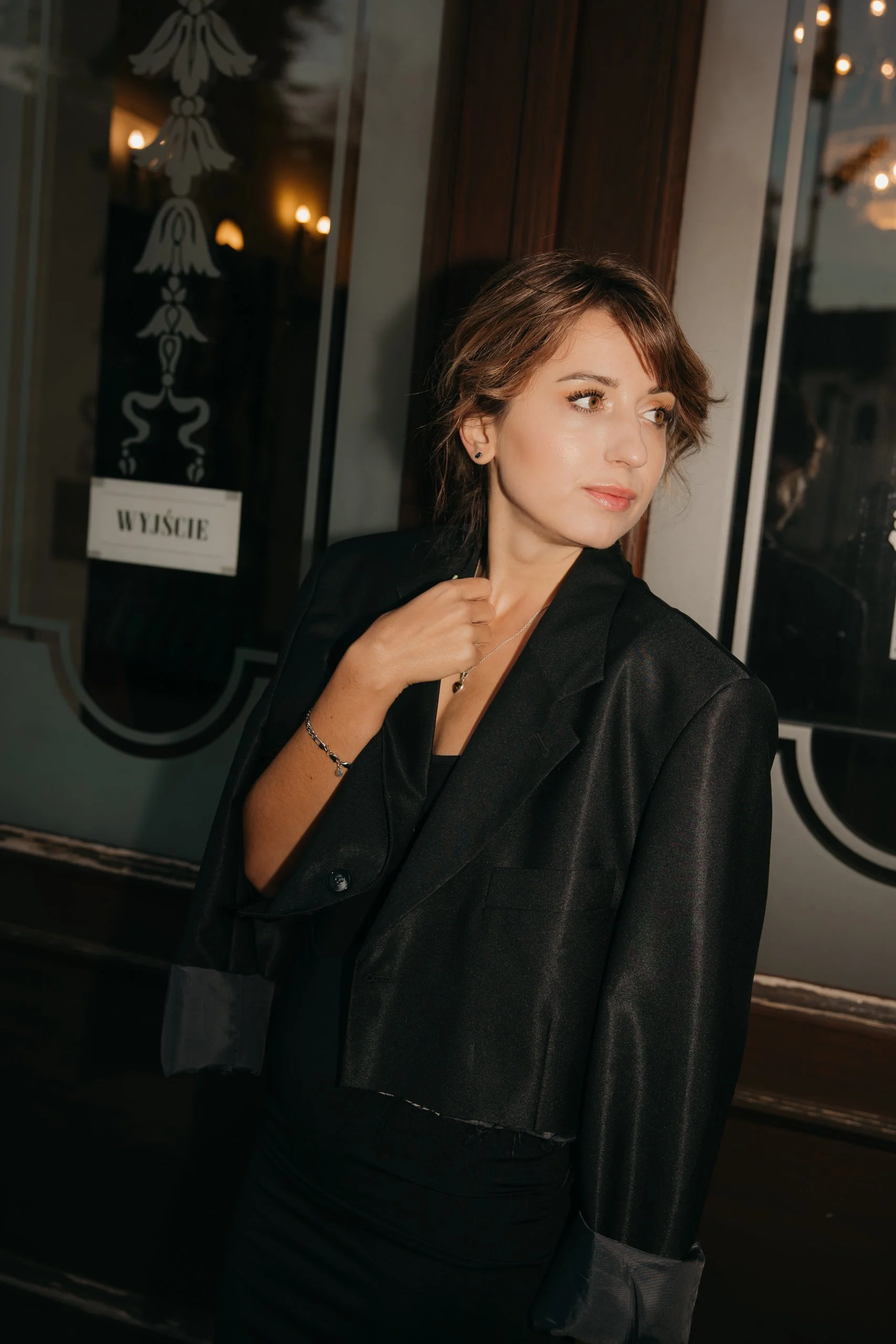 A woman with short brown hair, wearing a black blazer and dress, stands near a window with a reflective surface, looking to her right with a slight smile. There is a dark wooden wall behind her and a sign with the word 'WYJŚCIE' and an emblem on it, 
