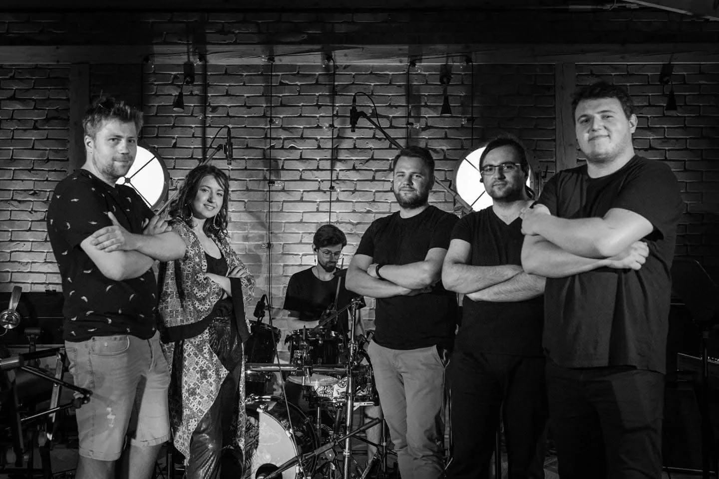 A black-and-white photo of a band with five members in a music studio with exposed brick walls. Four men are standing with crossed arms, and one man is sitting at a drum set in the background.