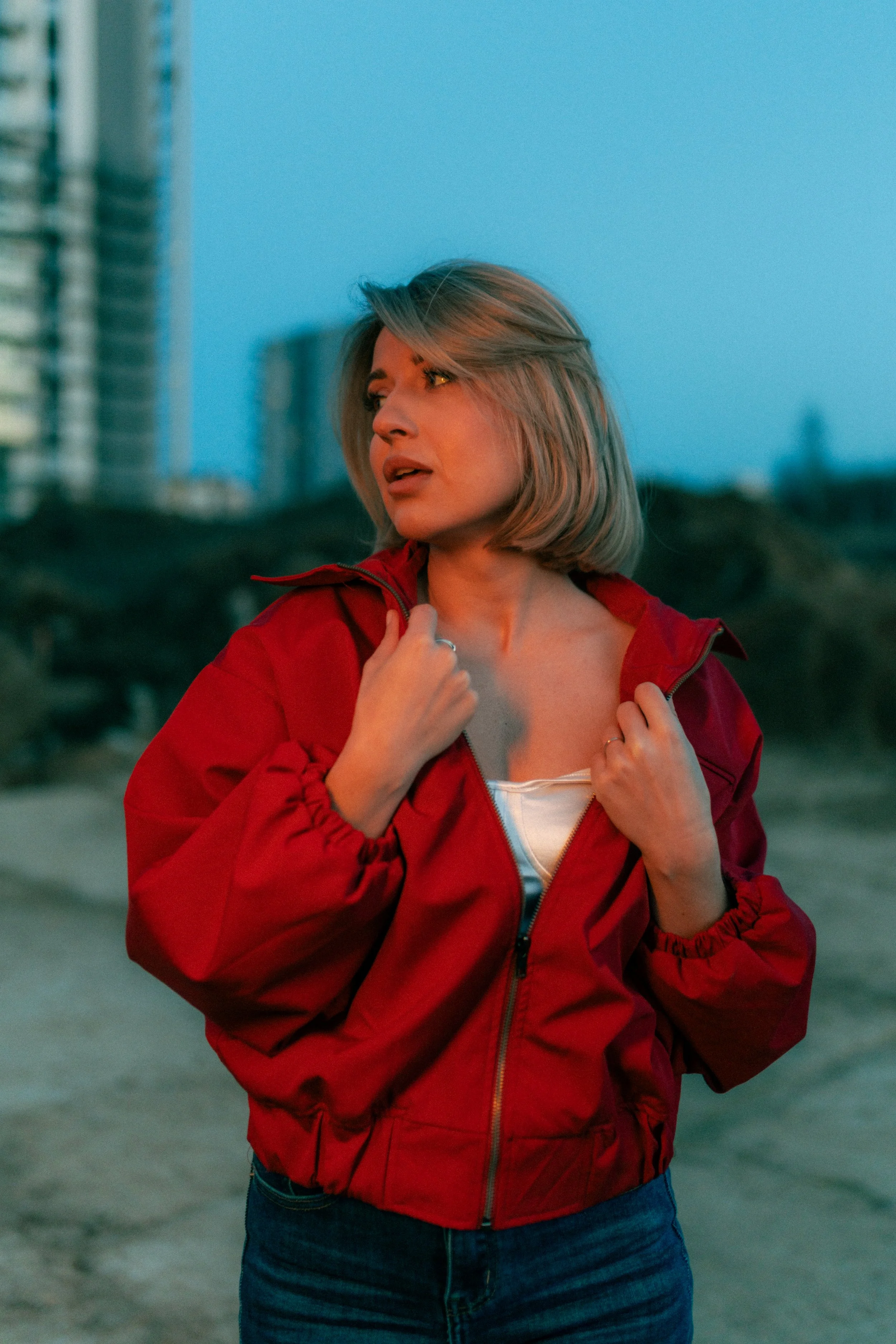 A woman with blonde hair wearing a red jacket and blue jeans, standing outdoors during dusk or early evening.