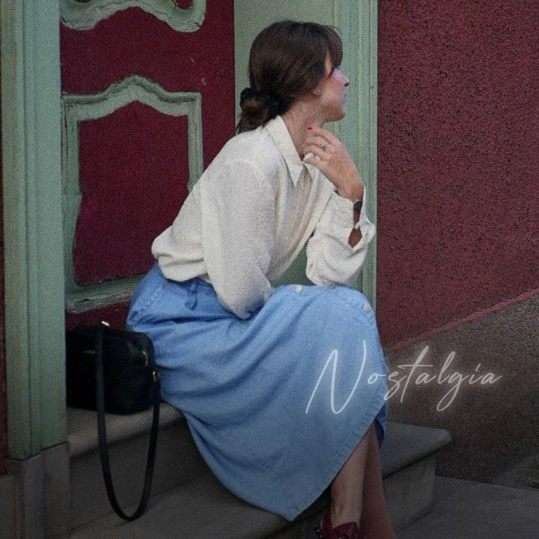 A woman sitting on a bench at a bus stop, dressed in a white blouse and a long light blue skirt, with a black bag beside her, looking to the side.