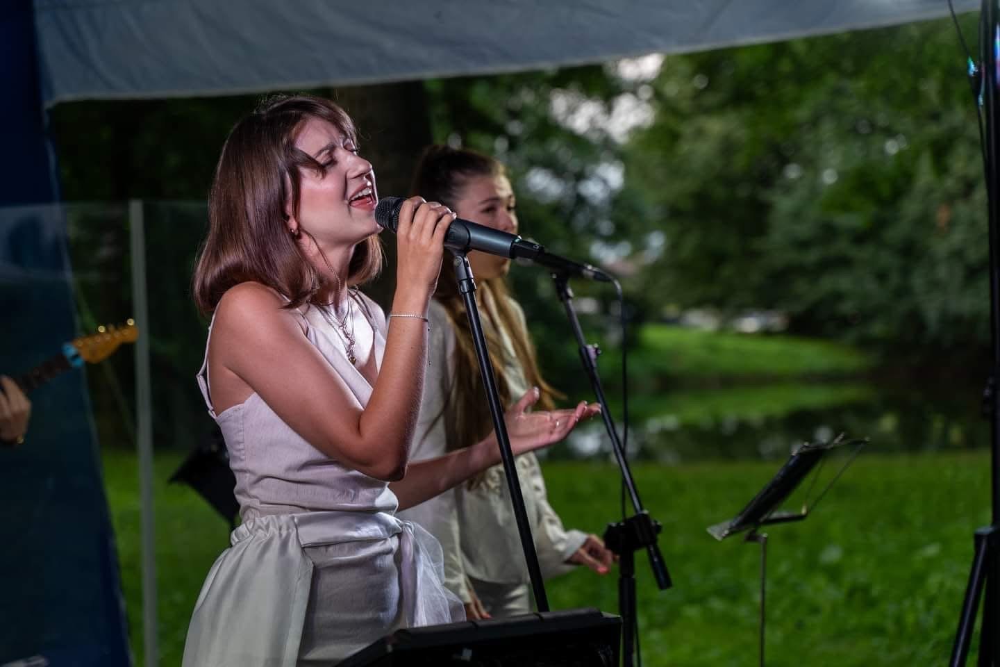 Two women singing into microphones during an outdoor music performance with trees and greenery in the background.