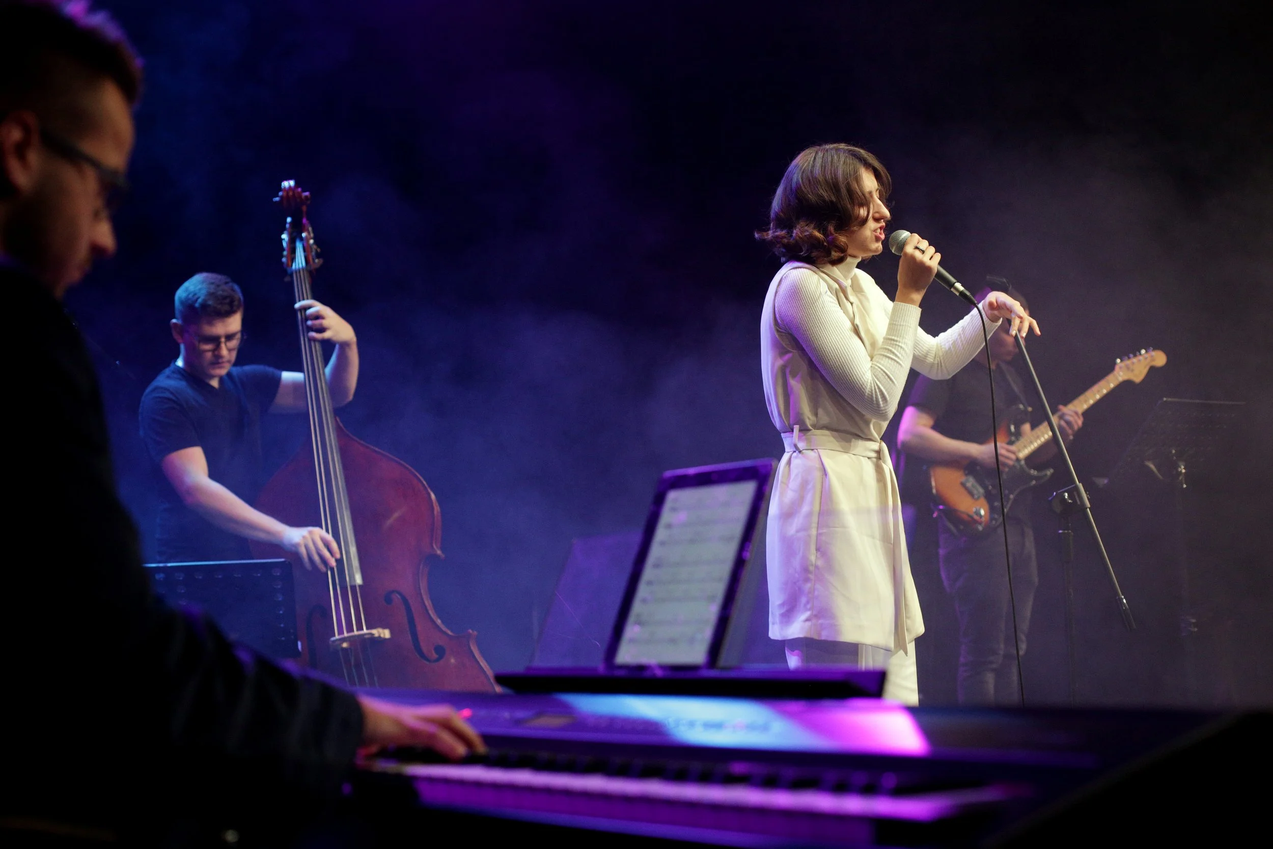 A group of musicians is performing on stage: a woman singing into a microphone, a man playing the electric guitar, a man playing the bass, and another person playing the keyboard.