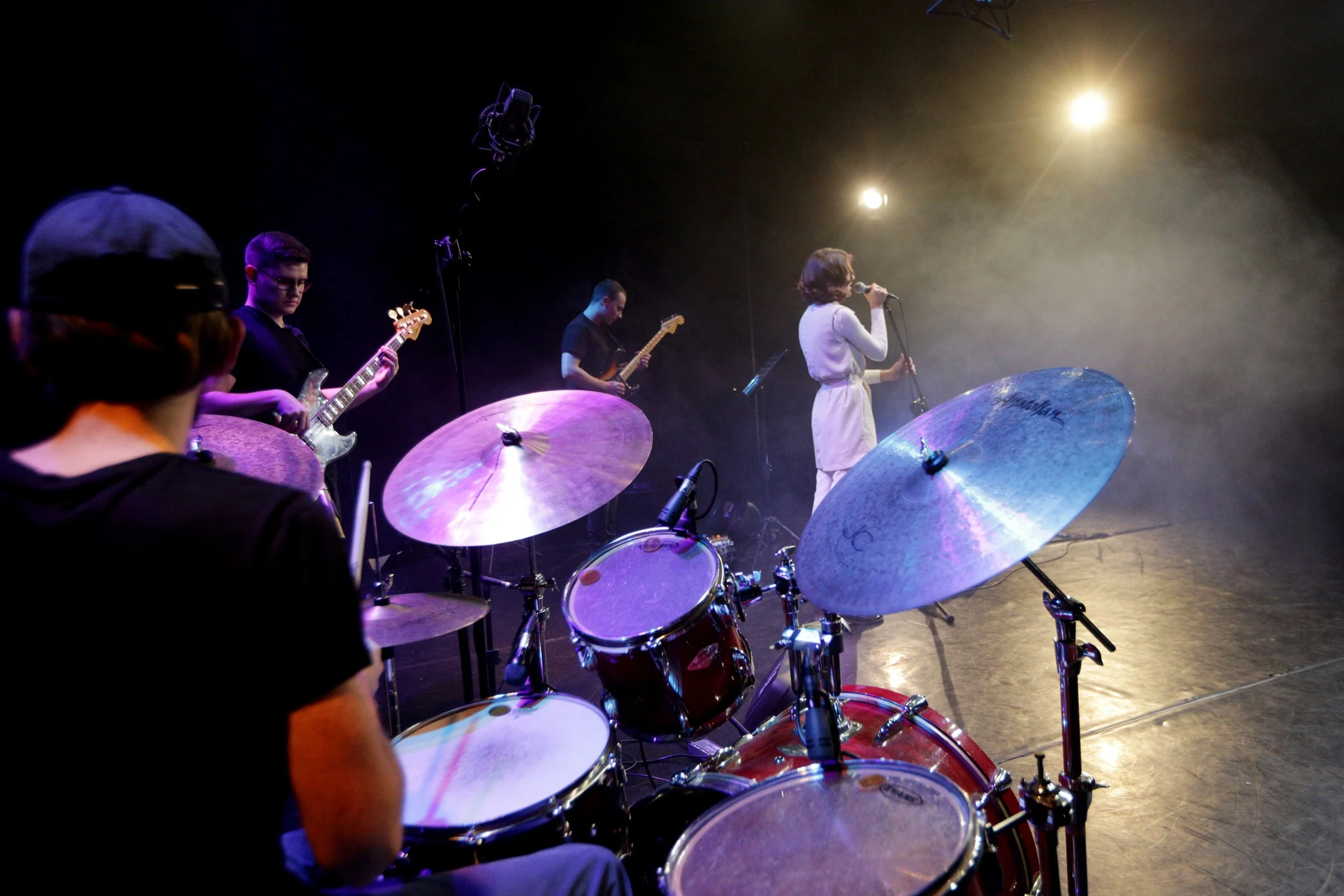 A band performing on stage with a drummer in the foreground, other musicians playing guitar, a vocalist singing into a microphone, stage lights shining down, and a smoky atmosphere.