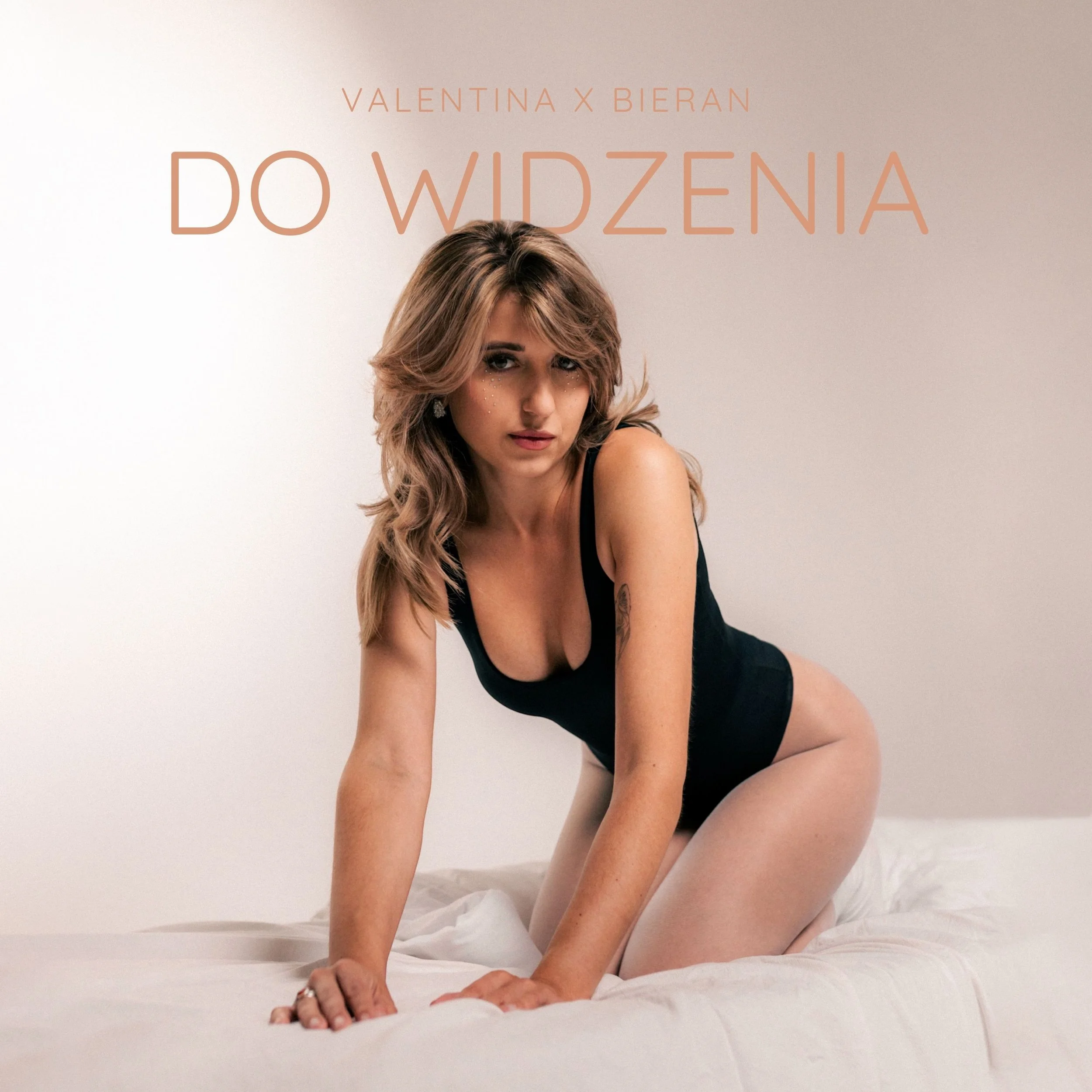 A woman poses on her hands and knees on a bed with white sheets. She has long, wavy, blonde hair and is wearing a black top with thin straps. Her face is turned slightly to the right, and she is looking at the camera with a neutral expression.