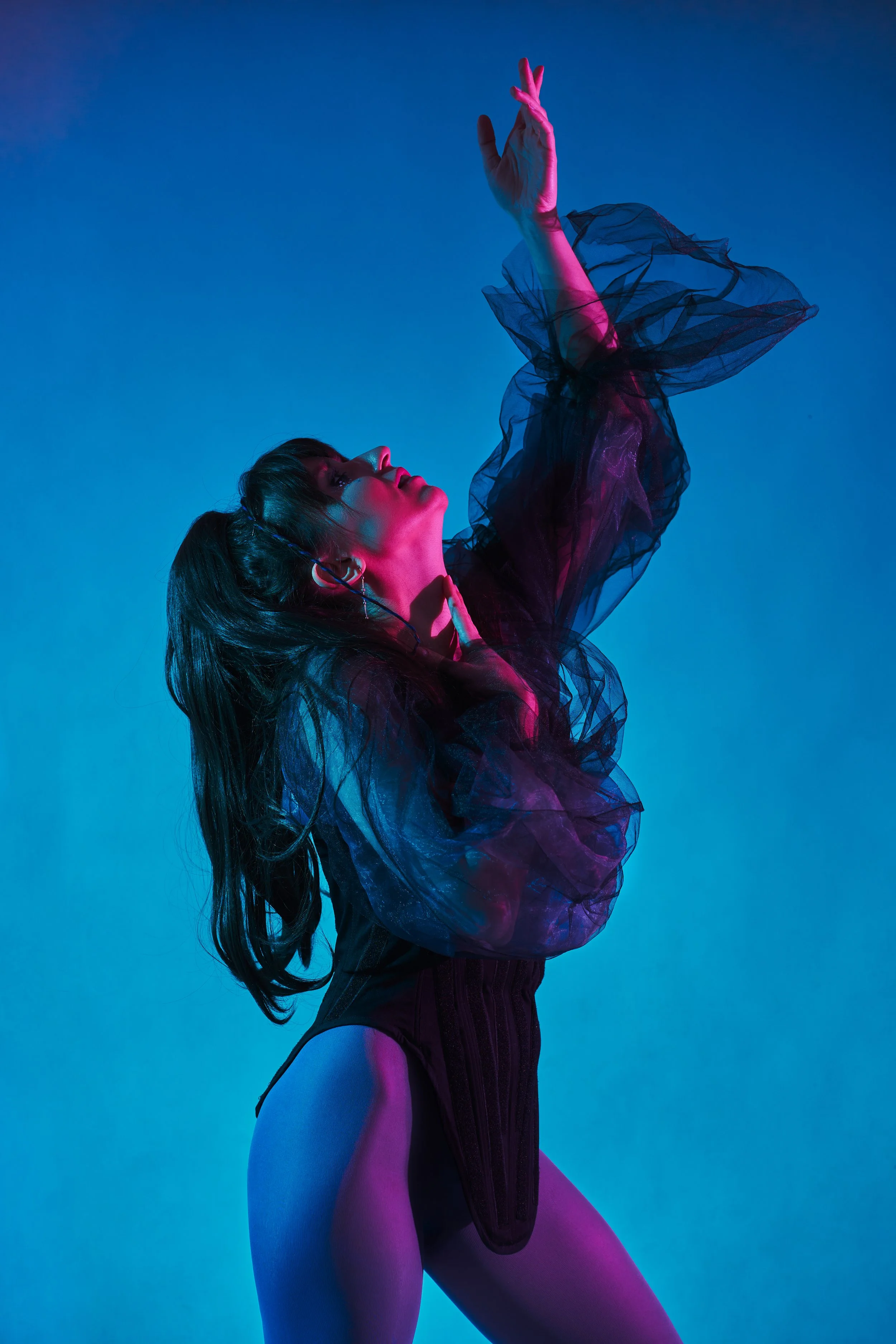 A woman dances in colorful, neon lighting against a blue background. She wears a sheer black top, high-waisted shorts, and stands in a dramatic pose with one hand raised above her head.