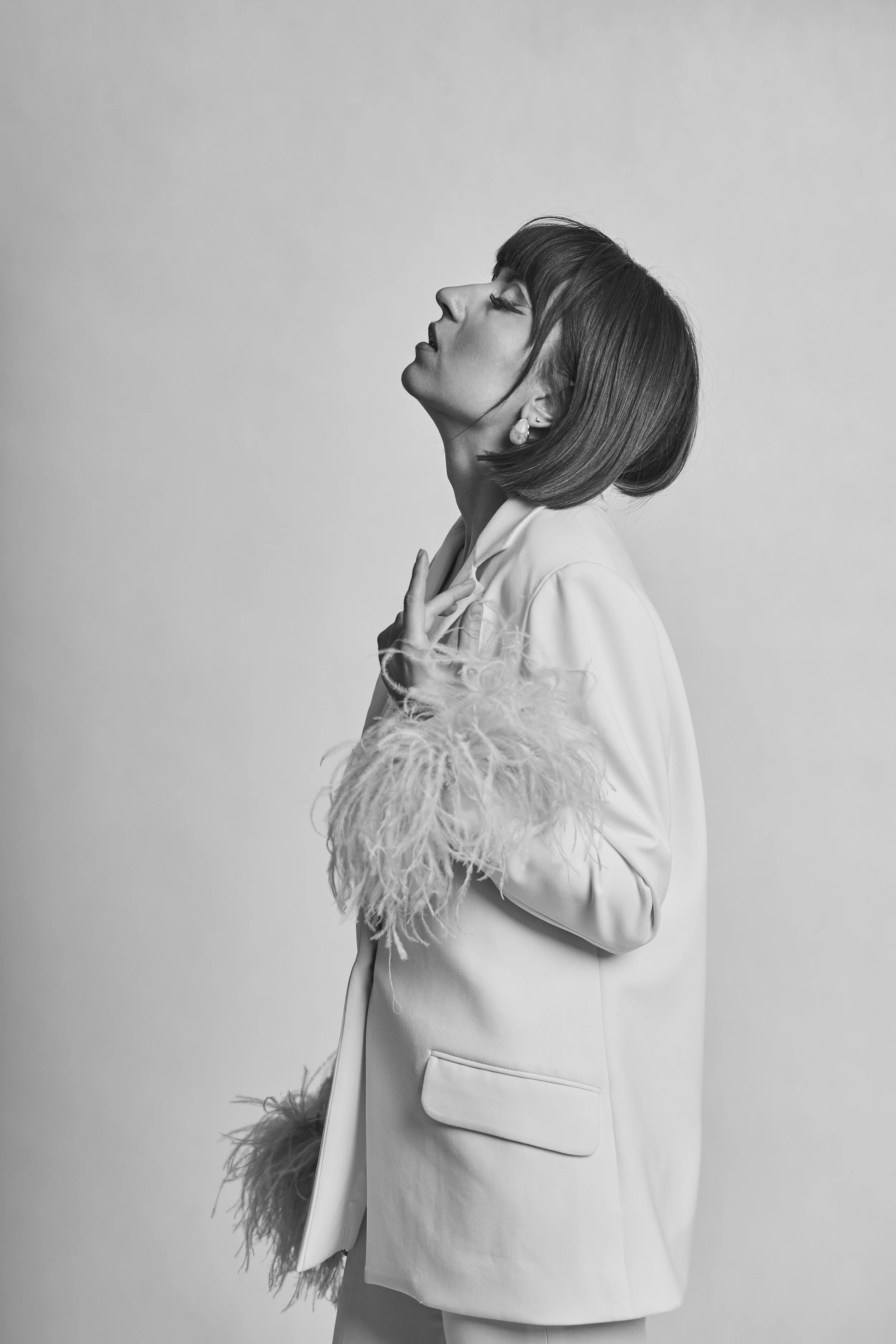 Black and white photo of a woman with short hair, dressed in a stylish white coat with feathered cuffs, posing with her head tilted back and eyes closed.