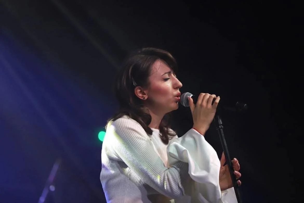 A woman singing into a microphone on stage with dark background and colorful stage lights.