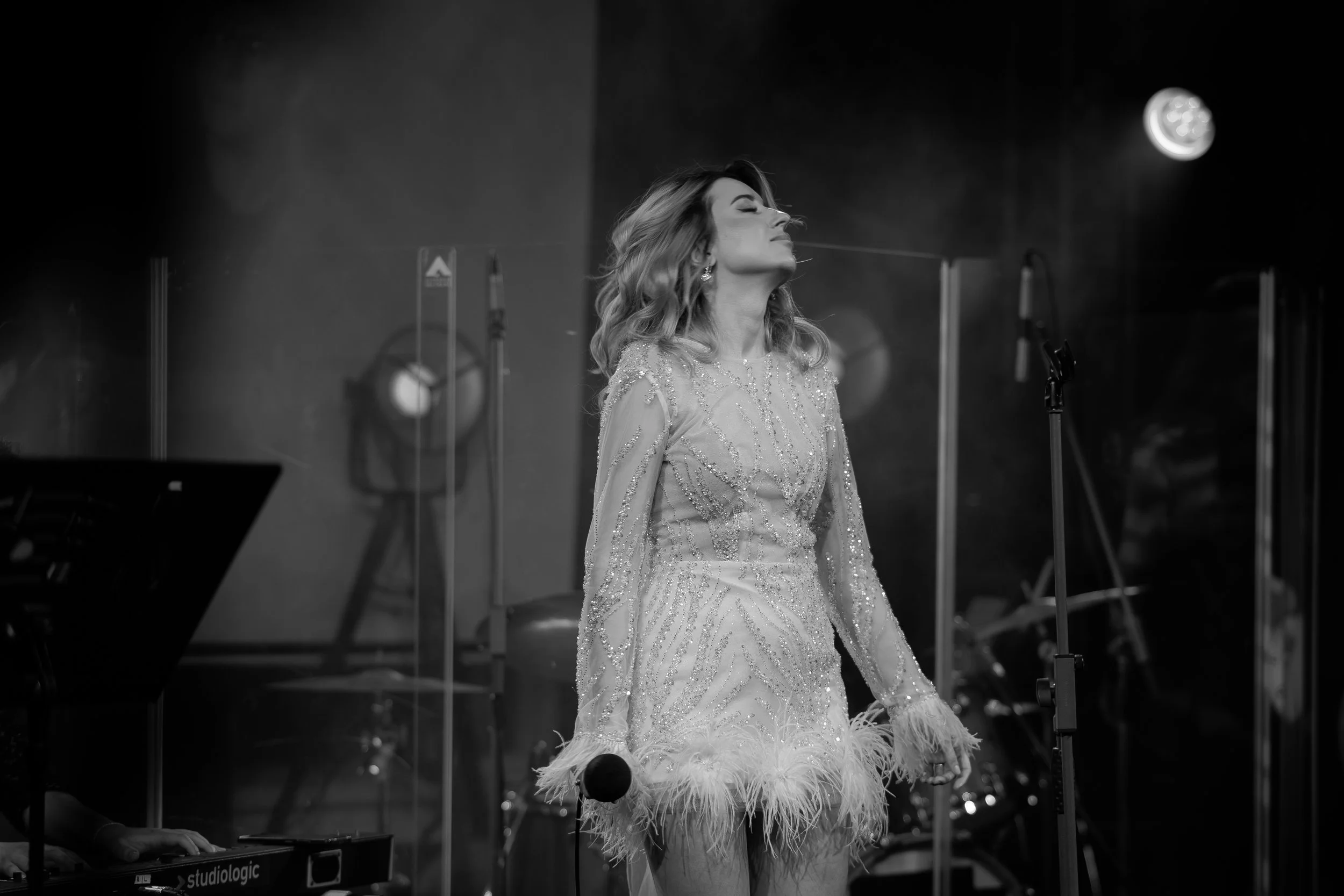 A woman with wavy hair wearing an ornate, sequined gown with feather embellishments at the bottom, standing on stage with eyes closed, surrounded by stage equipment and crew.