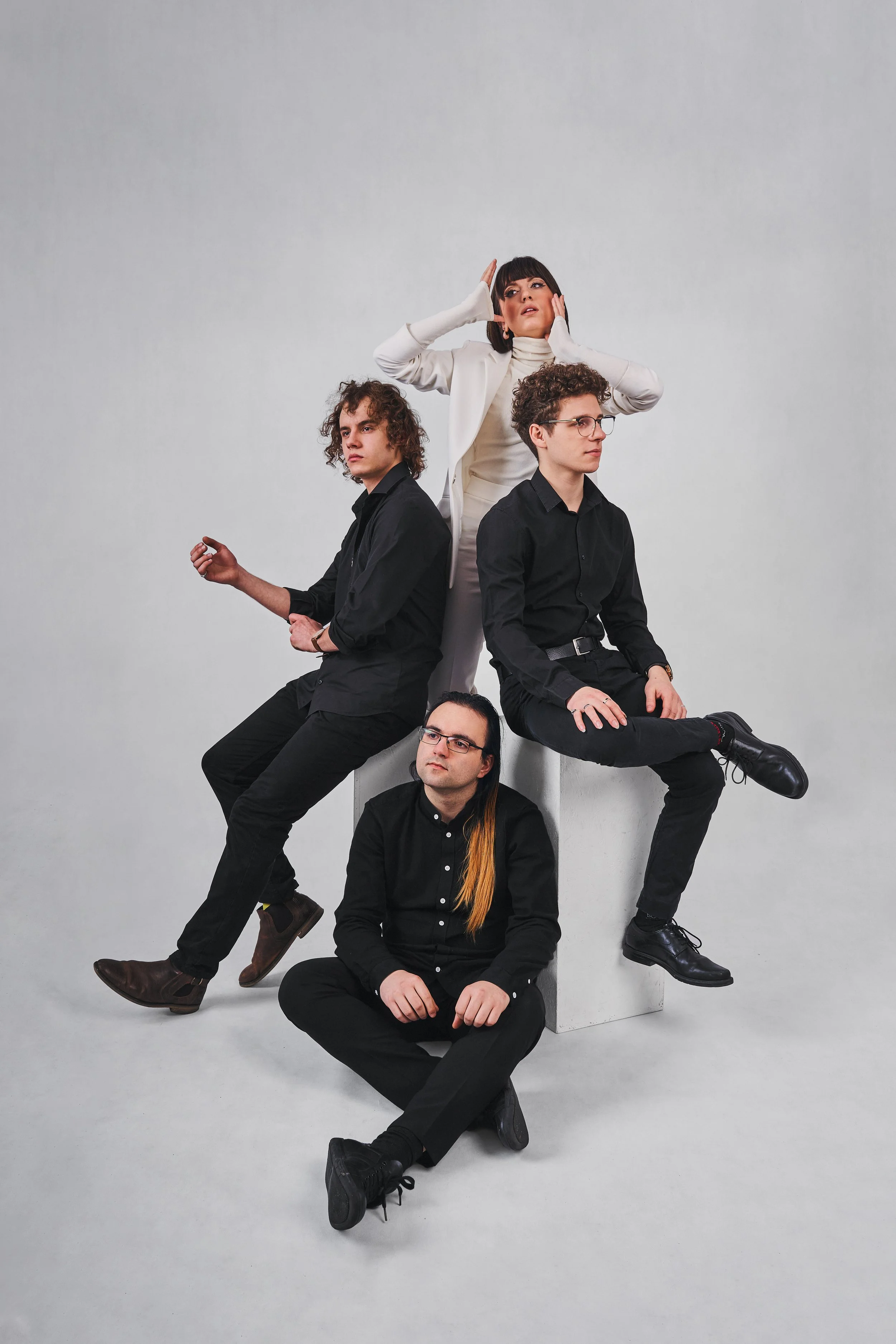 A group of five people arranged in a creative formation against a plain gray background. They are dressed in black and white clothing, with some standing, sitting, and lying down, and the central woman standing behind them with her hands on her face.
