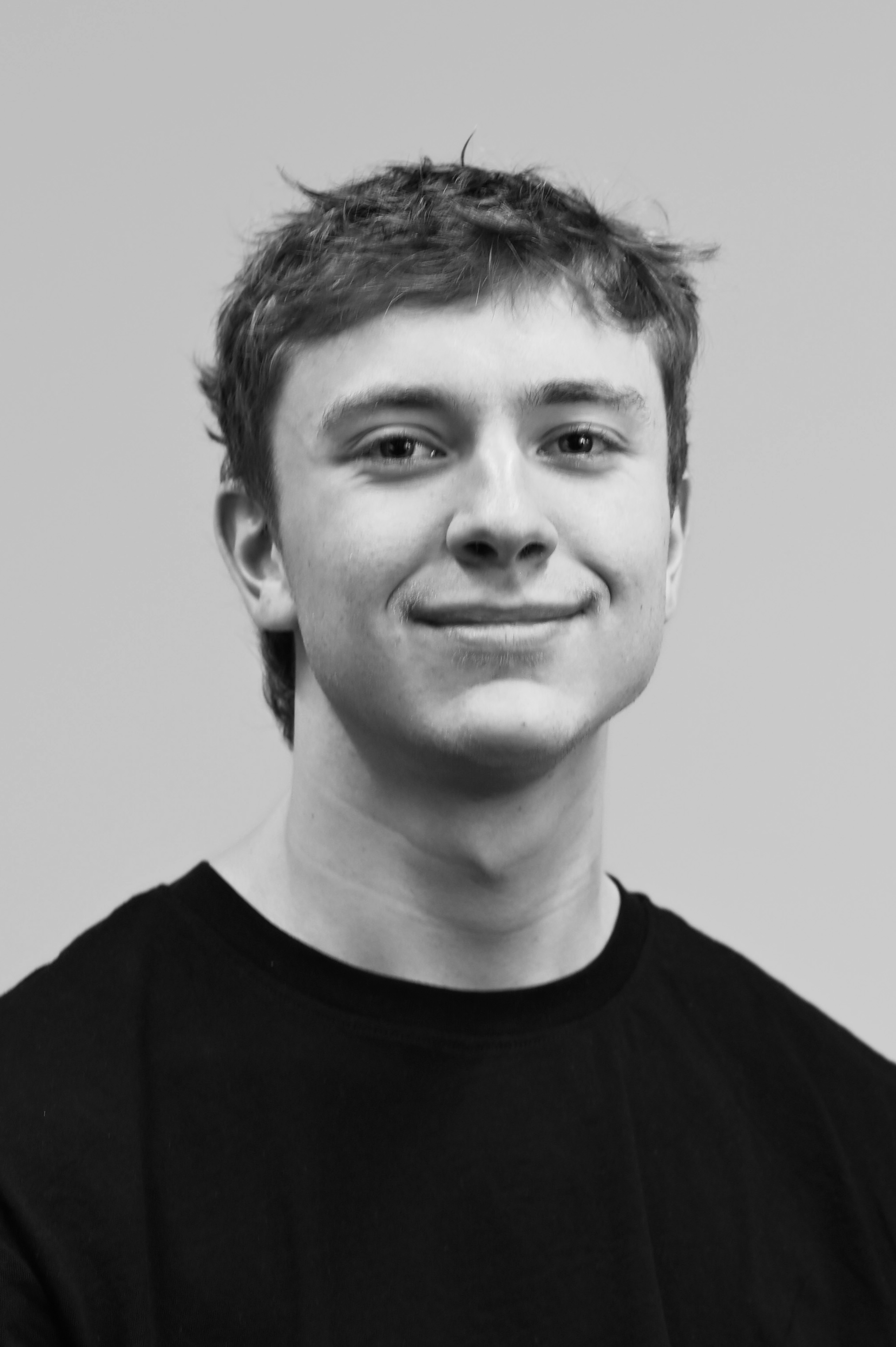Black and white photograph of a young man with short, slightly messy hair, wearing a black crew-neck shirt. He is smiling slightly and looking directly at the camera.
