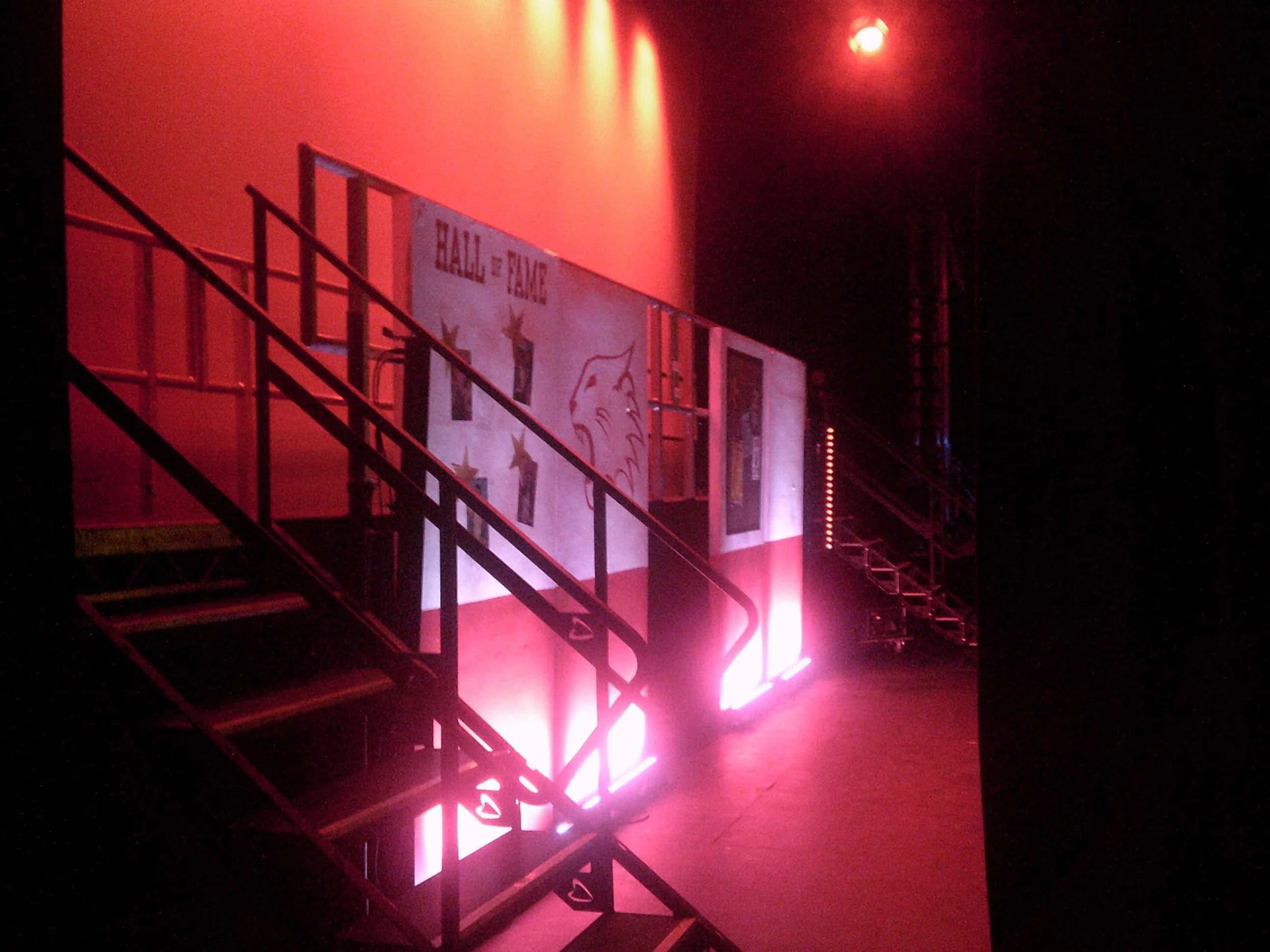 Stage with pink and purple lighting, metal stairs, and promotional posters for a talent show or event with stars and a cartoon face.