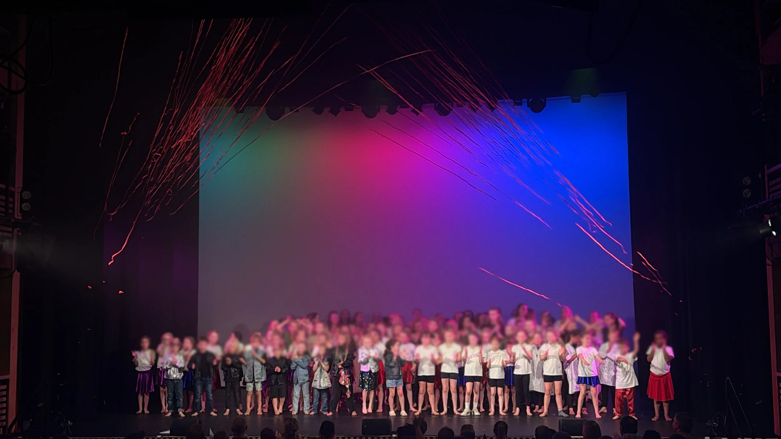 A stage with a large group of children standing in a line, under colorful lighting and confetti or streamers falling from above.