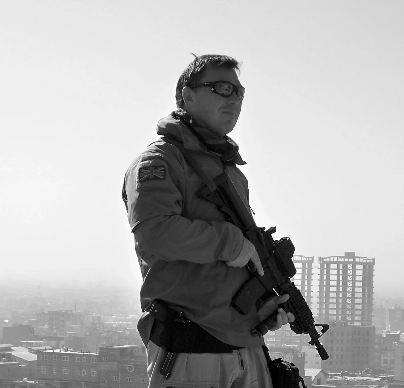 A man wearing sunglasses, a jacket with a UK flag patch, and holding a rifle, standing on a rooftop with city buildings in the background.