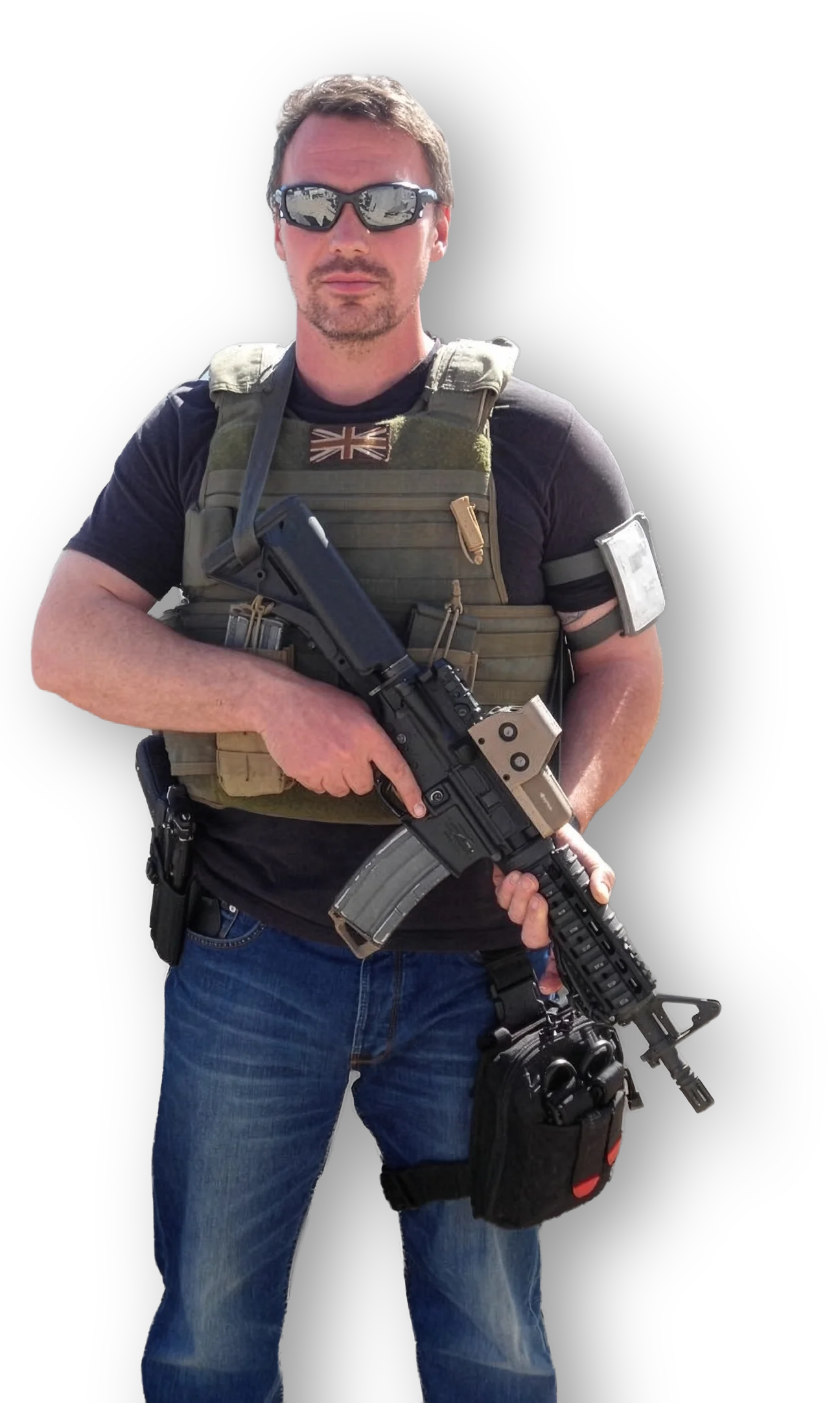 A man wearing sunglasses, a black T-shirt, and jeans, carrying a tactical vest and holding a rifle, with a bag attached to his waist, standing against a black background.