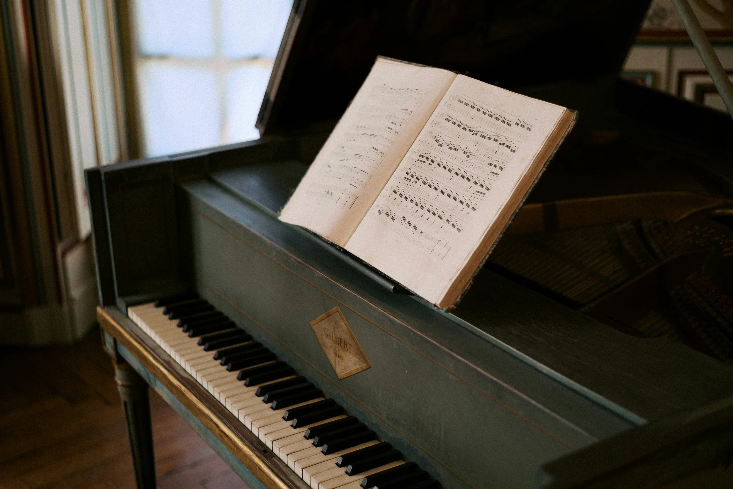 An antique upright piano with sheet music on the music stand, situated in a room with bookshelves and framed pictures in the background.