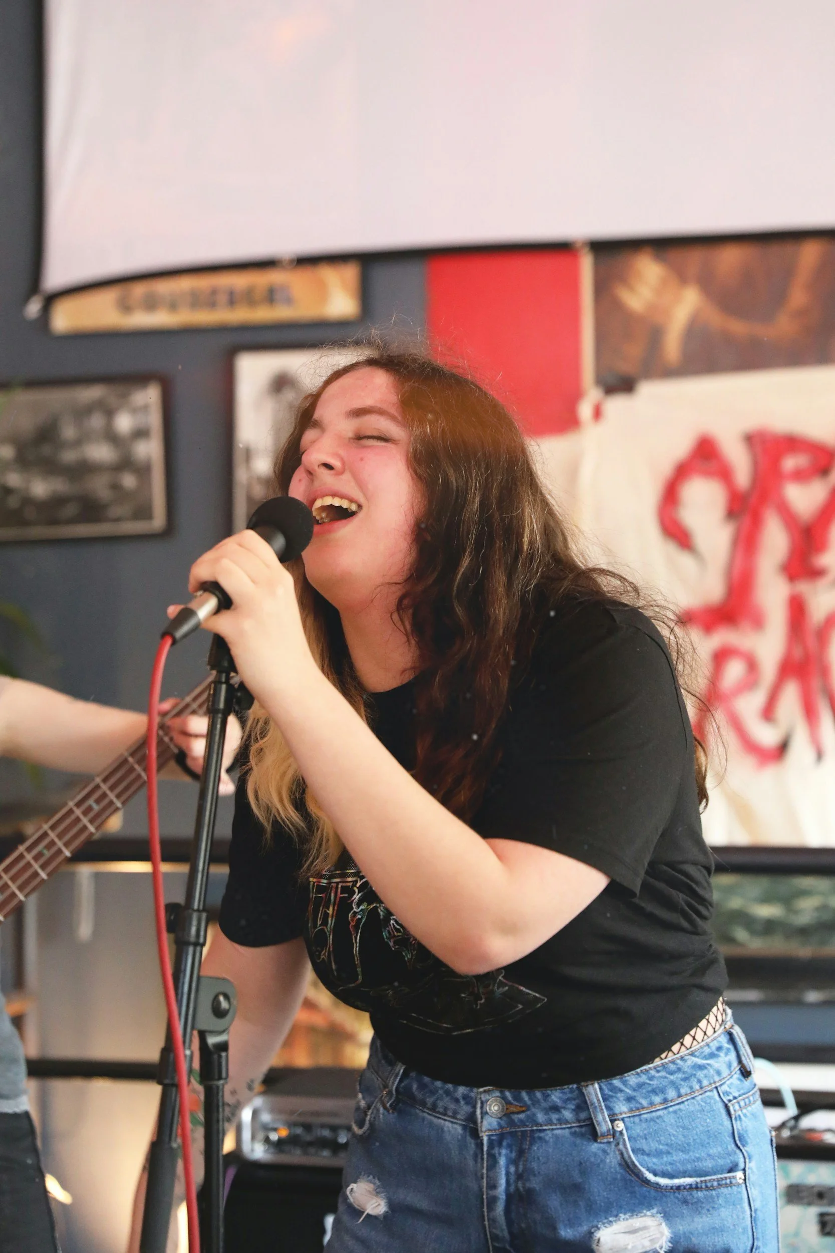 A young woman singing into a microphone with her eyes closed, wearing a black t-shirt and ripped jeans, in a room decorated with posters and pictures.