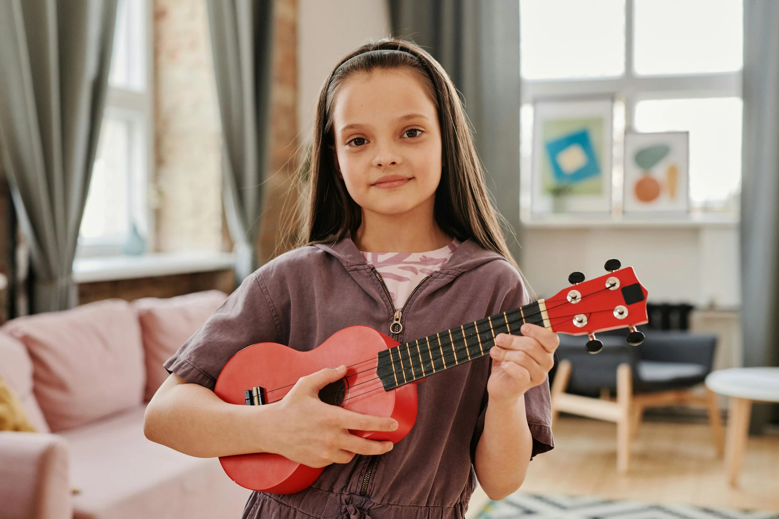 A young girl with long brown hair wearing a brown jacket and a pink and white striped shirt, playing a small red ukulele in a cozy living room with large windows, curtains, and colorful artwork in the background.