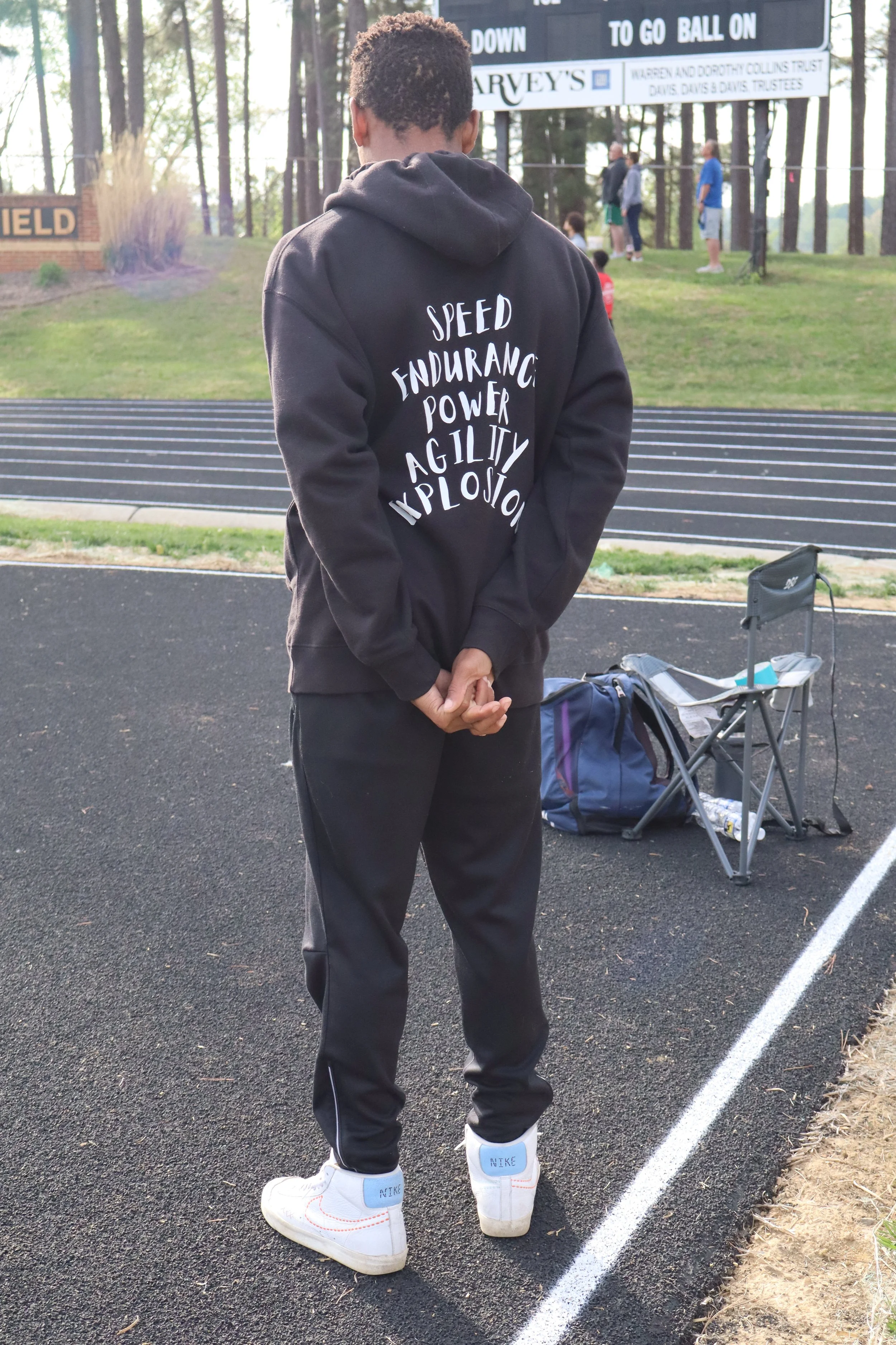 Person standing on a track at a sports field, wearing a black hoodie with white text and white Nike sneakers.