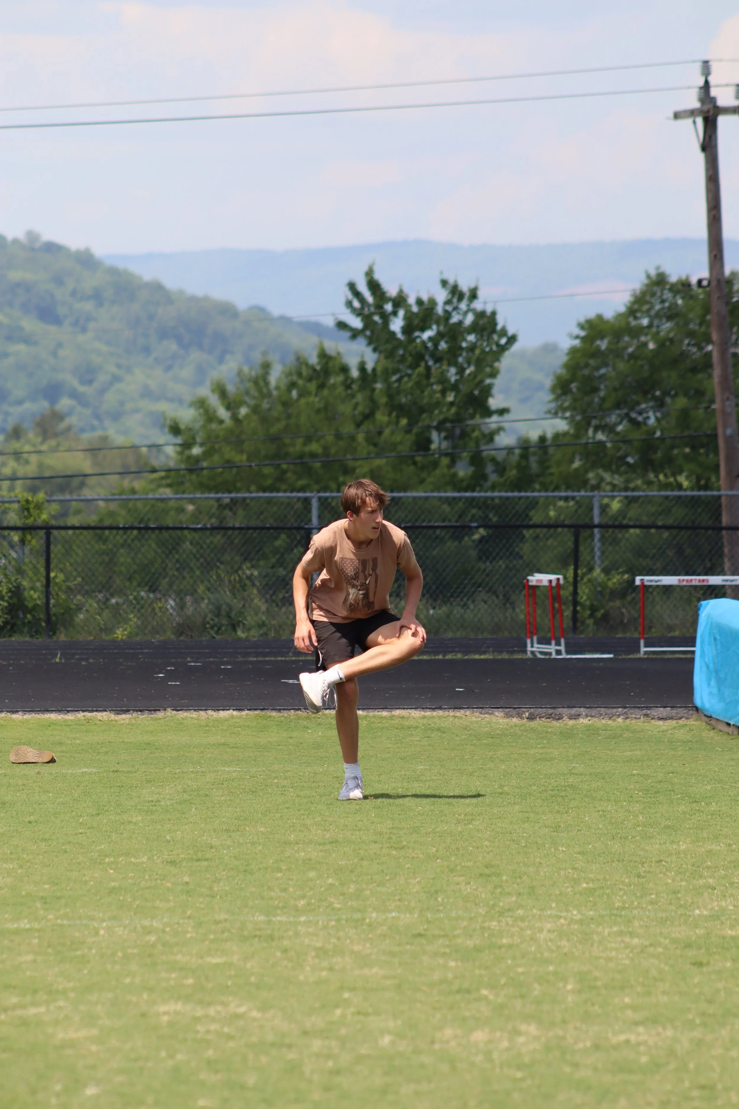 A young male athlete on a track field, preparing to kick a soccer ball, with hurdles, green grass, trees, and overcast sky in the background.