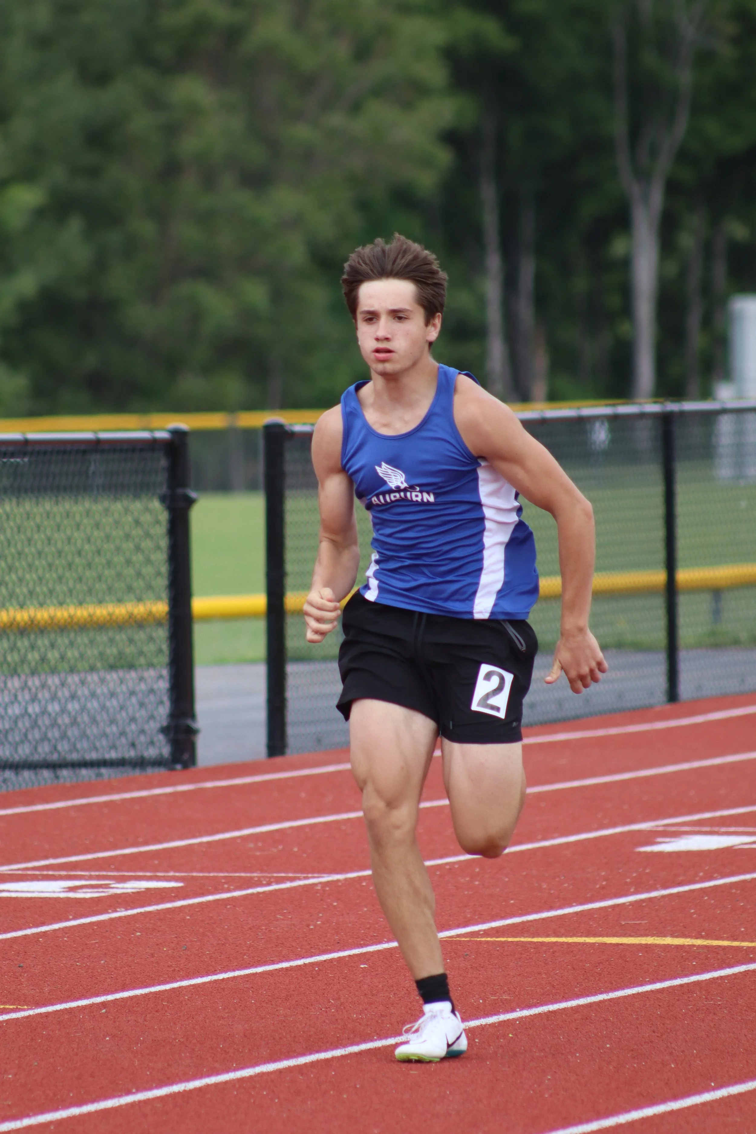 A young male athlete with brown hair running on an outdoor track, wearing a blue and white sleeveless jersey with a winged logo, black shorts with a number 2 bib, and white running shoes with neon green soles.
