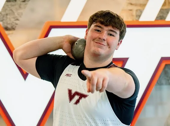 A young male athlete smiling and pointing at the camera, holding a shot put over his shoulder, wearing a Virginia Techsports jersey in a gym or indoor sports facility.