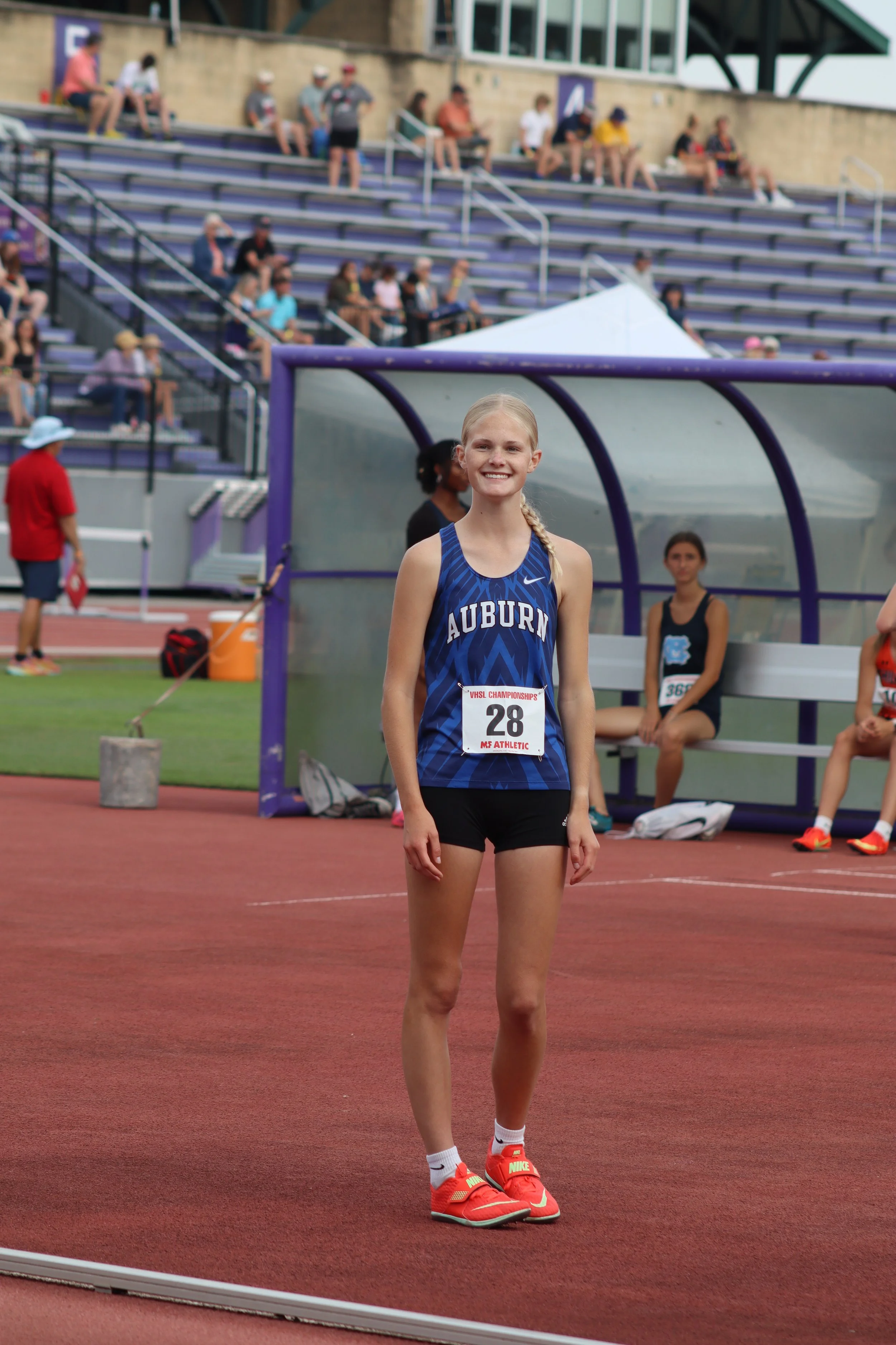 A young female athlete with blonde hair in a braid, smiling, wearing a blue Auburn track uniform with bib number 28, red Nike running shoes with neon green Nike logo, standing on a red track at a stadium during a race event.