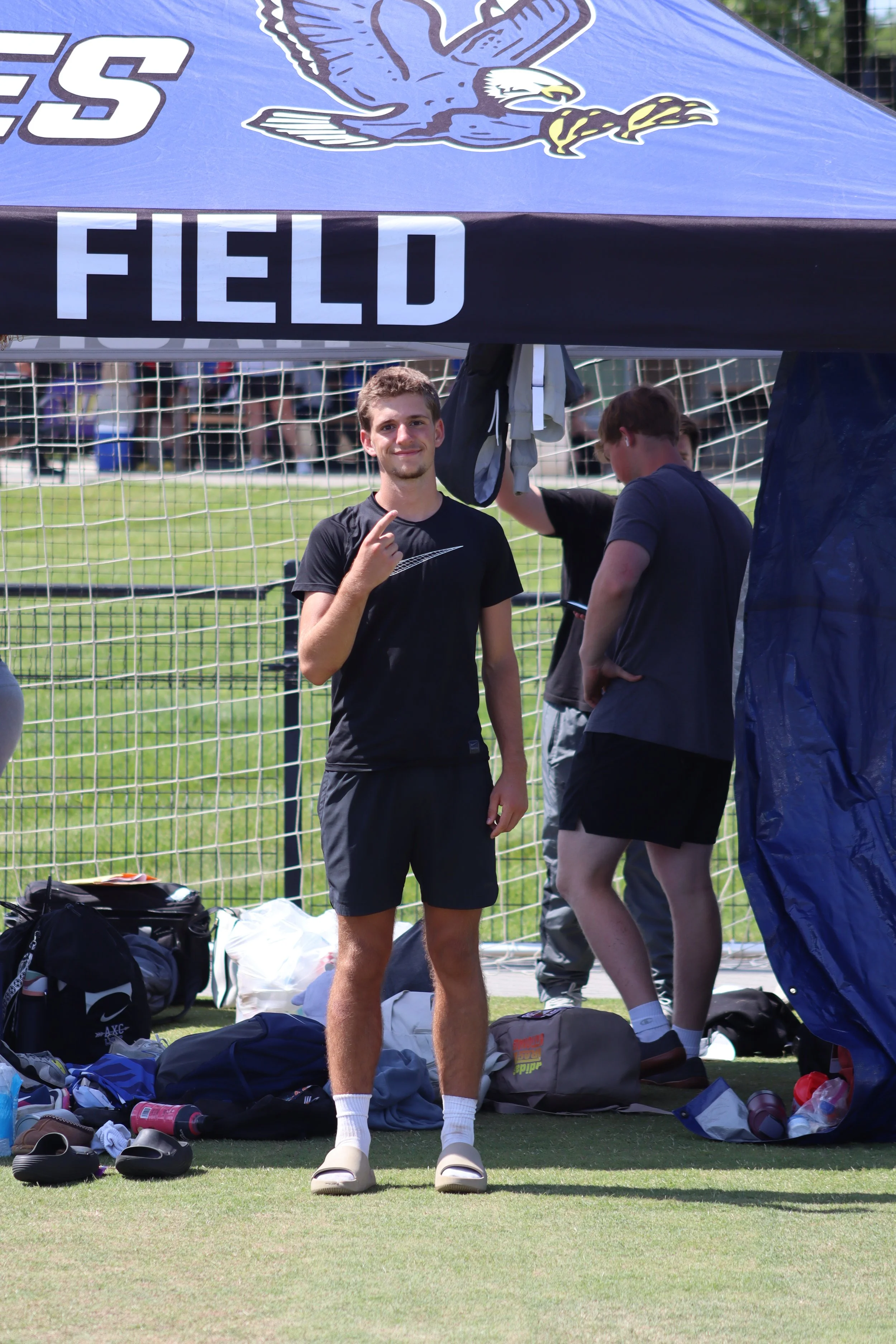 A young man standing on a soccer field in front of a tent that says "FIELD" with a logo of an eagle on it. He is smiling and pointing to himself. There are soccer shoes, bags, and other gear on the ground.