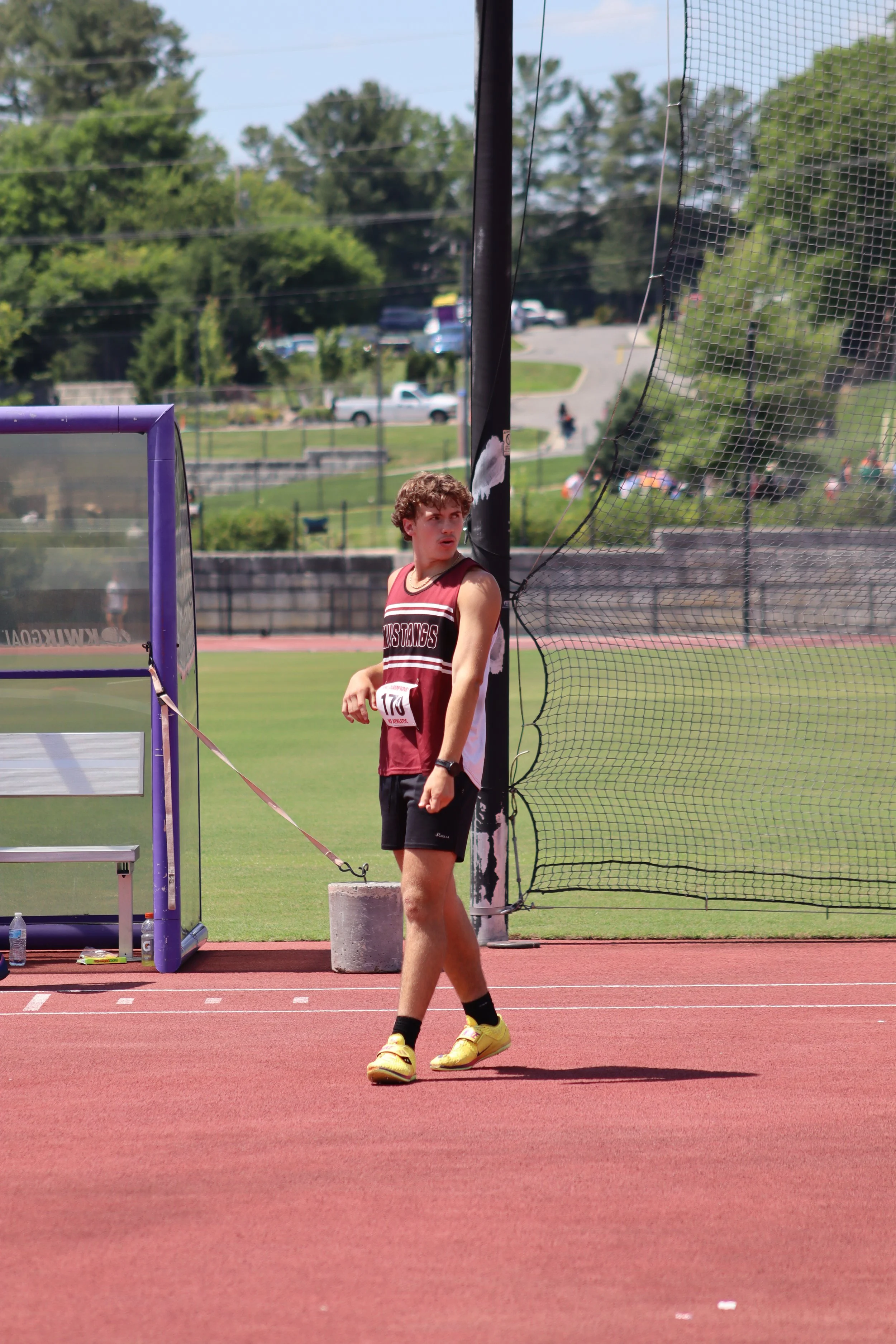 A teenage boy in a red and black sleeveless sports uniform standing on a red running track near a high jump or pole vault area at an outdoor track and field venue. There is a netting structure nearby and greenery in the background.