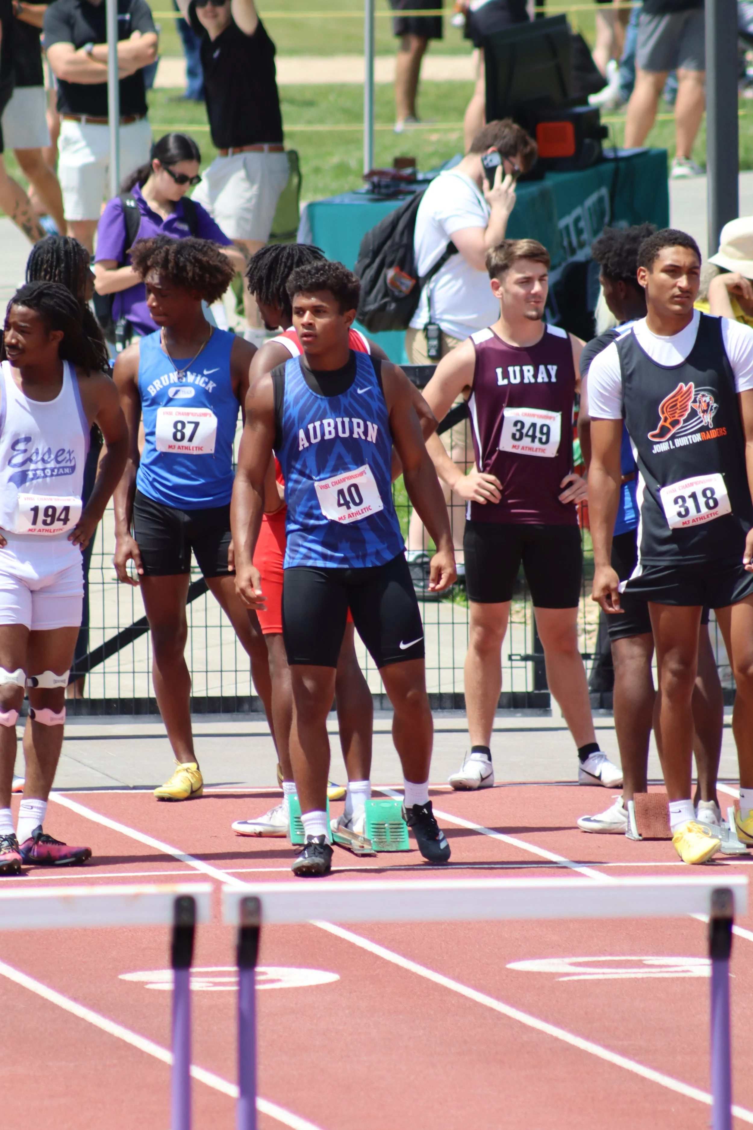 A group of young athletes at a track and field event, preparing to start a race, with hurdles set on the track and spectators and officials in the background.