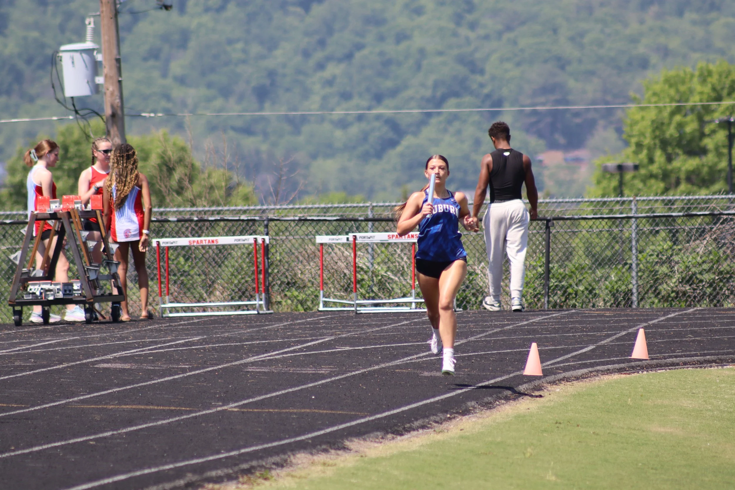 A young female athlete running on an outdoor track while holding a relay baton. There are four women in sports attire at a table on the sidelines, with hurdles and cones set up on the track. The background features green trees and a mountain range under a clear sky.