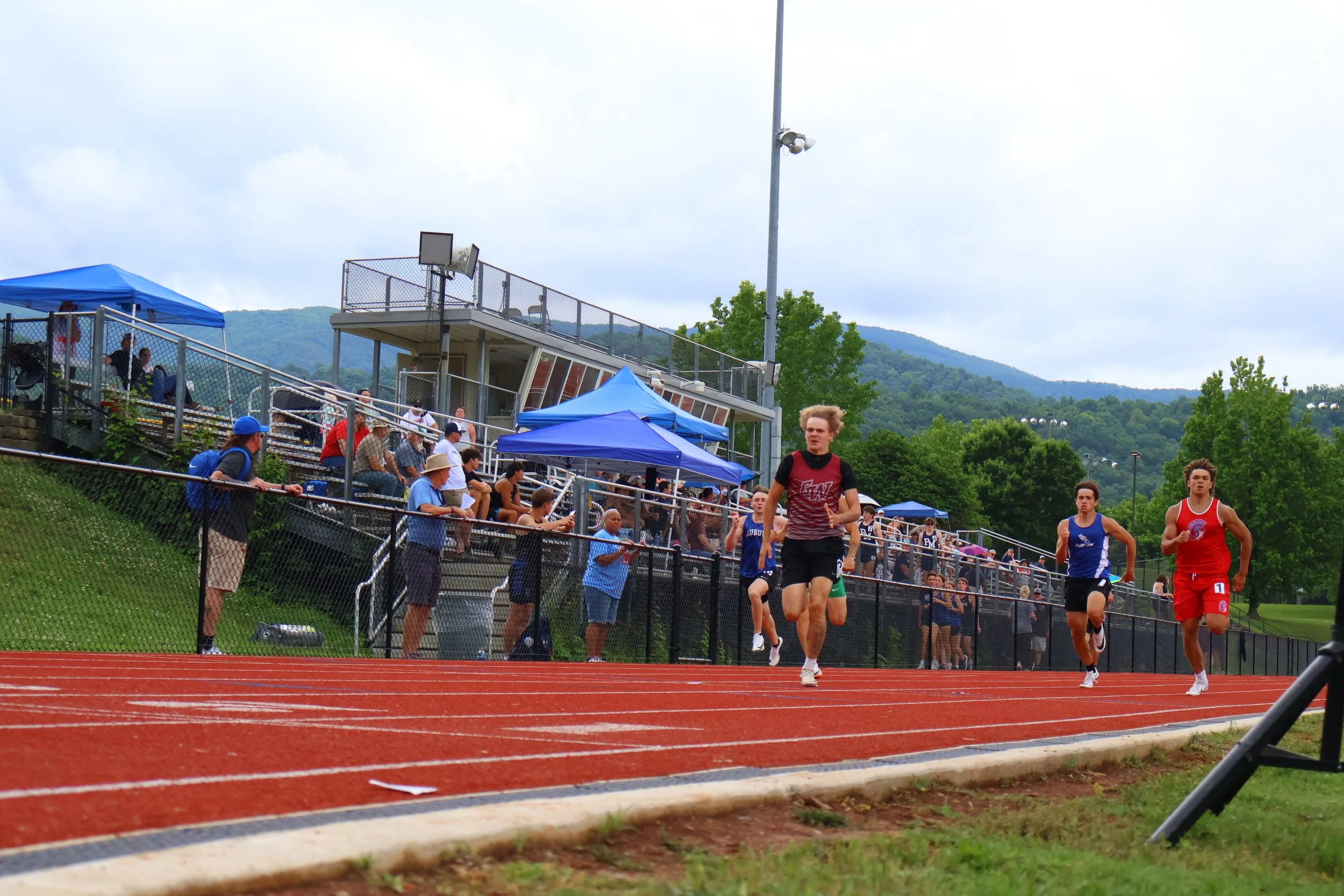 A track and field event at an outdoor stadium during daytime, with runners in red and blue uniforms competing on a red running track. Spectators and coaches sit on bleachers covered with blue tents, with a hilly landscape and cloudy sky in the background.