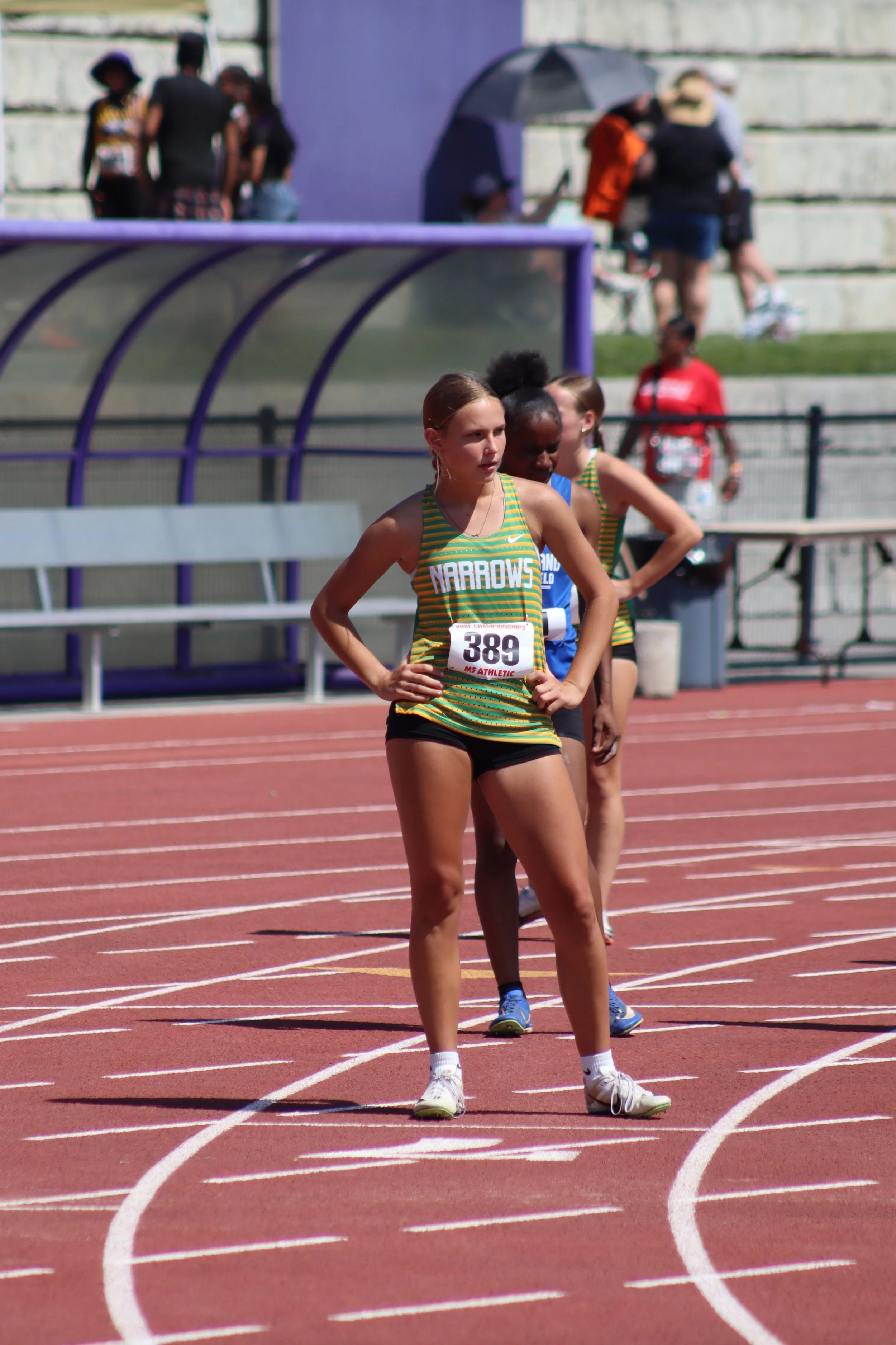 Female athletes in green and yellow uniforms with black shorts standing on a red running track, preparing for a race with a stadium in the background.