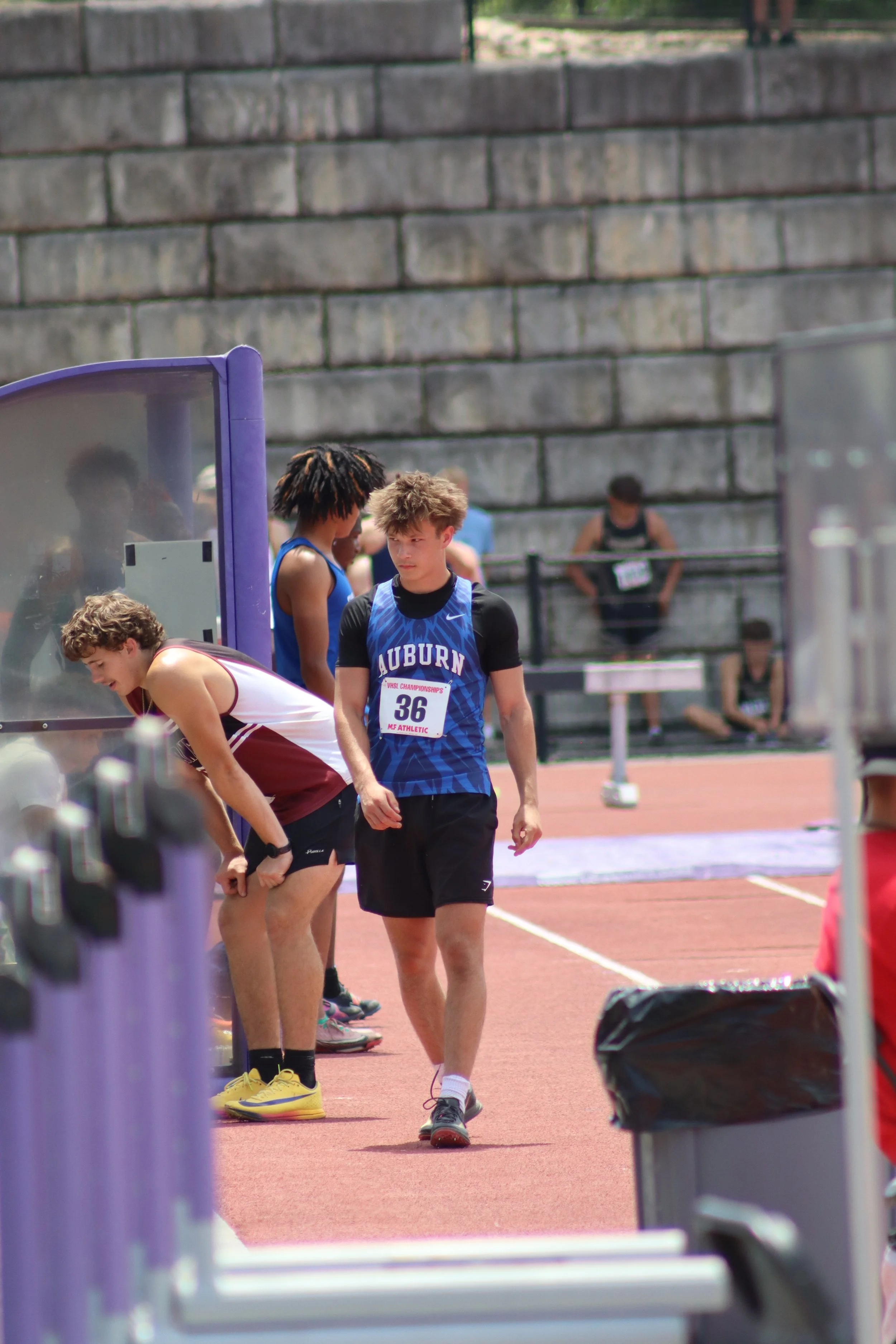 Young male athletes preparing for a track and field event on an outdoor track, standing near a purple hurdle with a stone wall in the background.