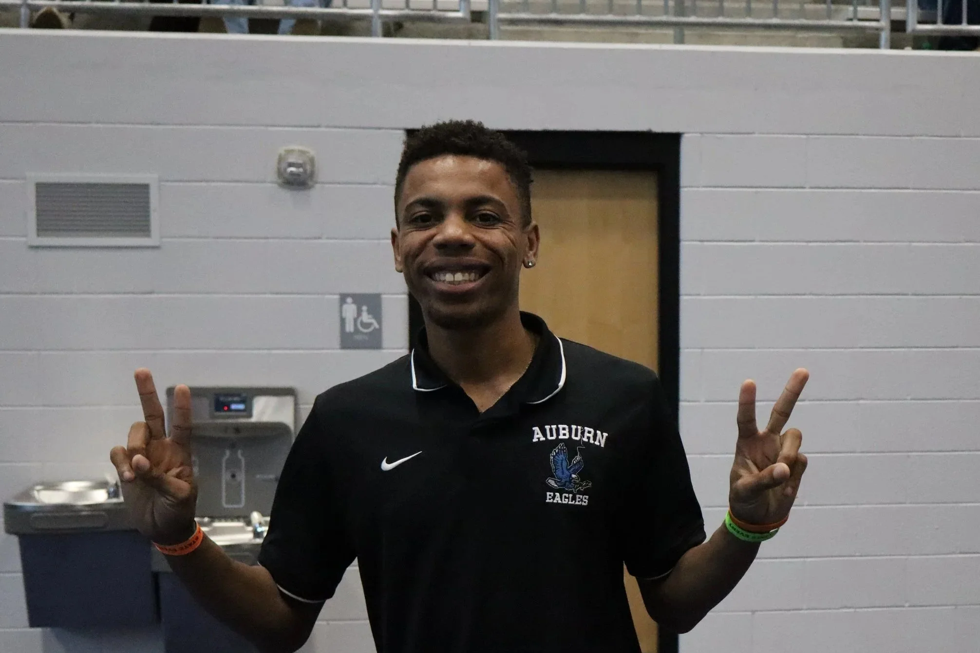 Young man smiling and showing peace signs with both hands, wearing a black Auburn Eagles shirt, standing in front of a light gray wall with a door and a sign indicating accessible restroom.