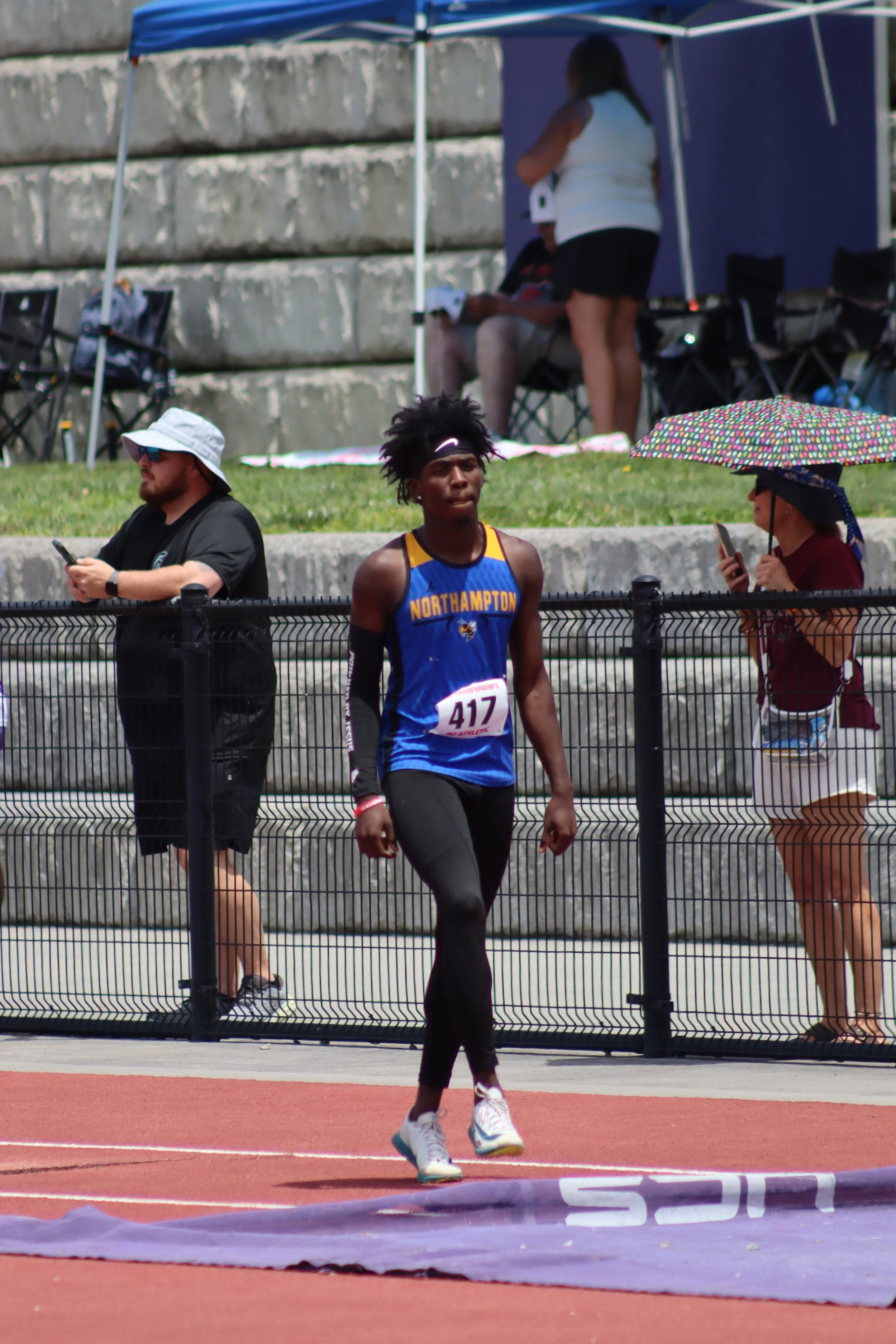 A male athlete wearing a blue and yellow jersey with the number 417 on it, walking on a track with a red surface during a sporting event. In the background, there are spectators, some sitting under shade tents, and a woman holding a colorful umbrella also using a mobile phone. A black fence separates the track from the spectators.