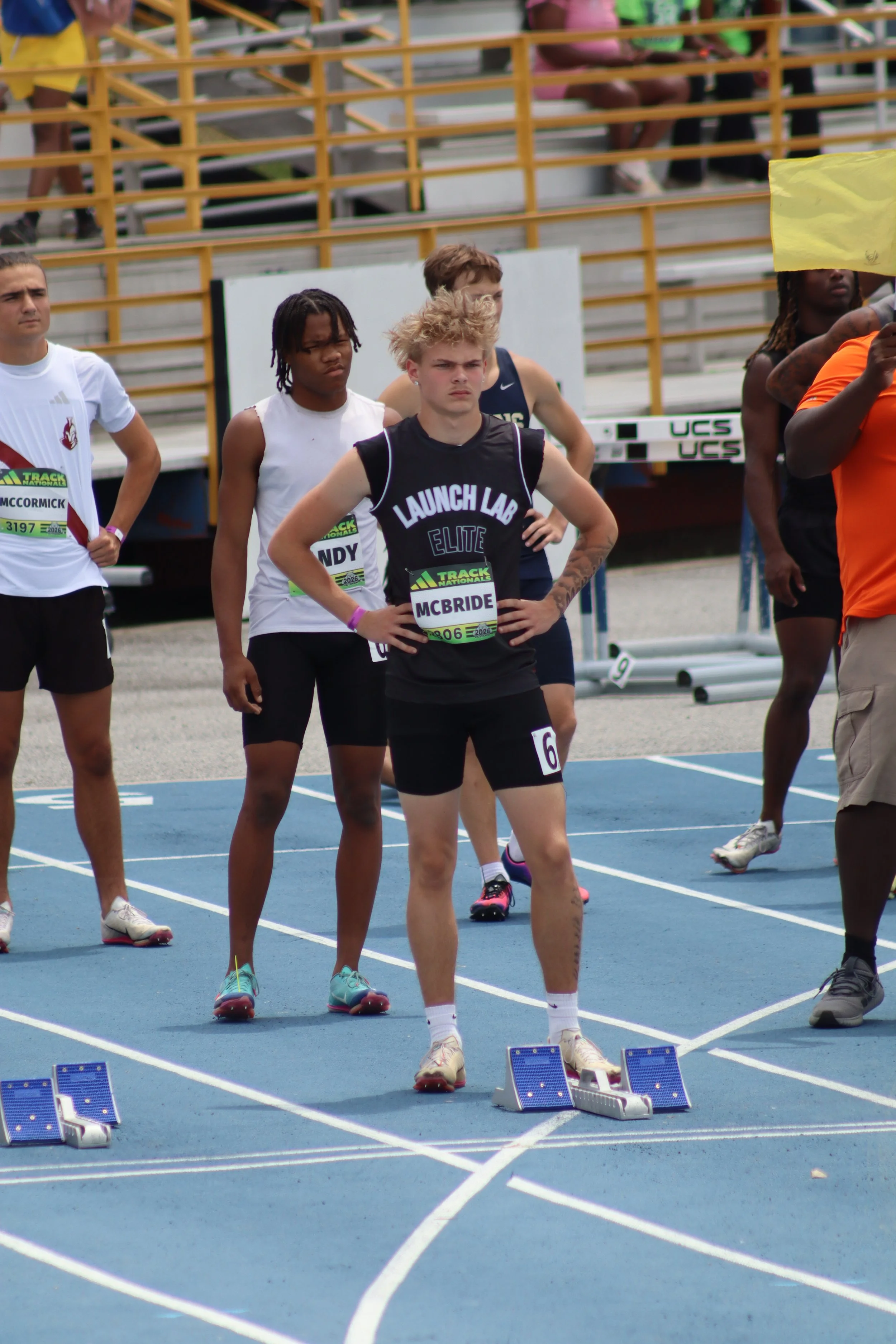 A track and field event at an outdoor stadium during daytime, with runners in red and blue uniforms competing on a red running track. Spectators and coaches sit on bleachers covered with blue tents, with a hilly landscape and cloudy sky in the background.