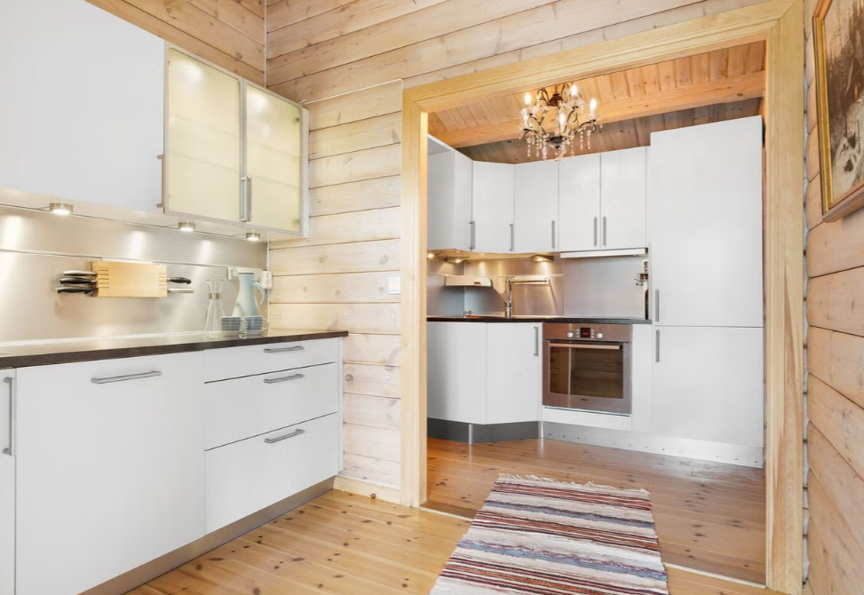 Kitchen with white cabinets, wooden walls, and a wooden floor, decorated with a chandelier and a colorful striped rug.