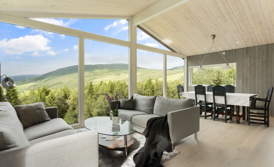 Living room with large glass windows showing a view of green mountains and trees, with two sofas, a glass coffee table, a dining table with six chairs, and wooden ceiling and walls.